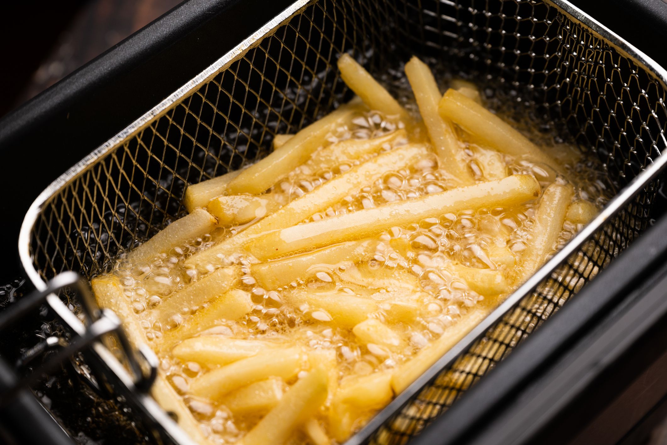 Closeup of basket with sticks of potatoes frying in oil to make french fries in a fryer, at a diagonal