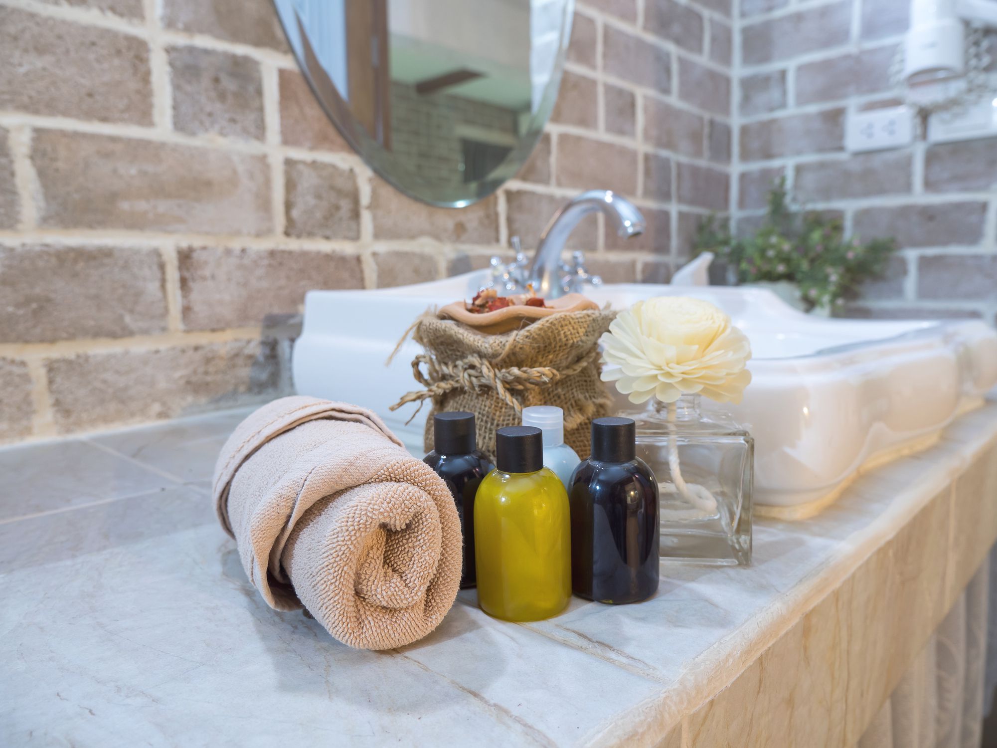 Focus on 4 vials of liquid soaps, a roll of natural towel, with a few other spa items on a counter with a sink in a bathroom with a brick wall and a mirror in the background