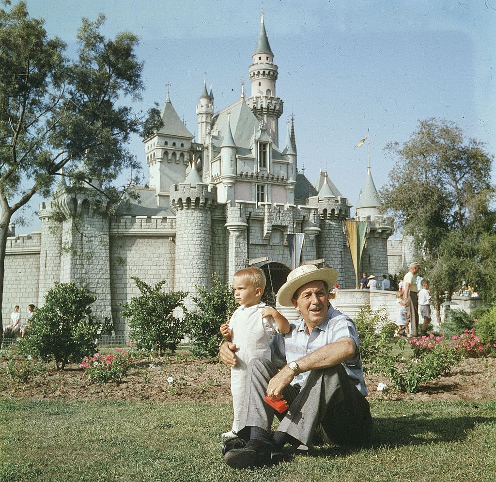 American animator and film studio founder Walt Disney (1901-1966) sits on a grassy lawn with his grandson, in front of the Magic Kingdom's castle at Disneyland, Anaheim, California