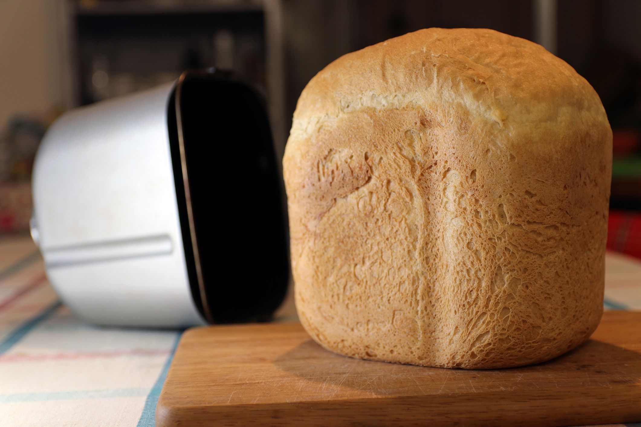 Selective focus of finished bread on a wooden cutting board with the bread maker pan, empty, in the background, blurred background