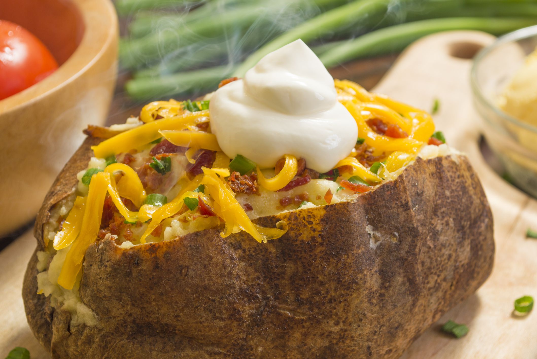 fresh hot baked potato with butter bacon cheese and chives topped with sour cream on a cutting board