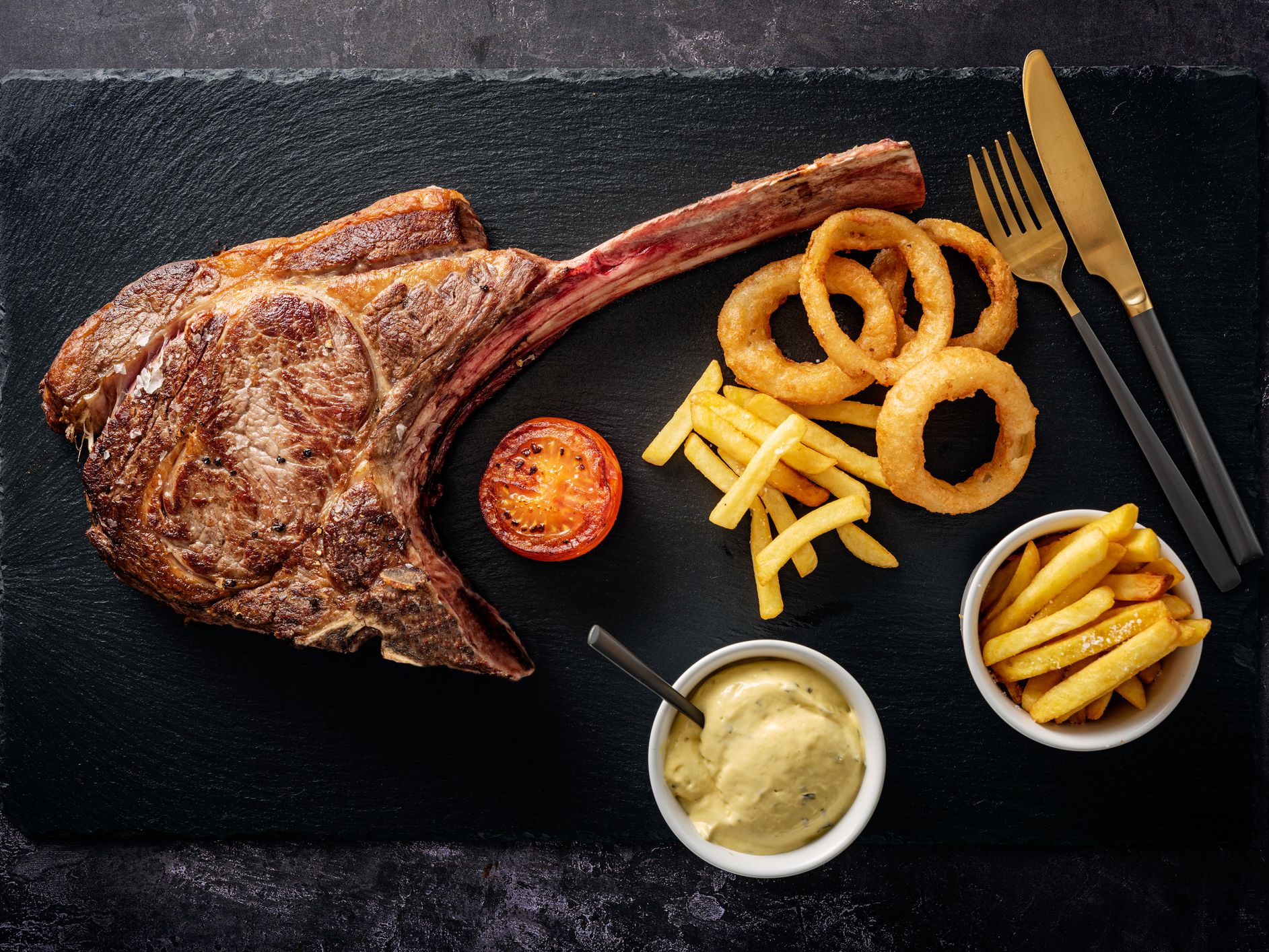 Overhead view of a huge, cooked tomahawk steak being served with; french fries, tomato slice, deep fried onion rings and béarnaise sauce served on a piece of oiled slate. Colour, horizontal with some copy space. The Tomahawk Steak got it’s name from their
