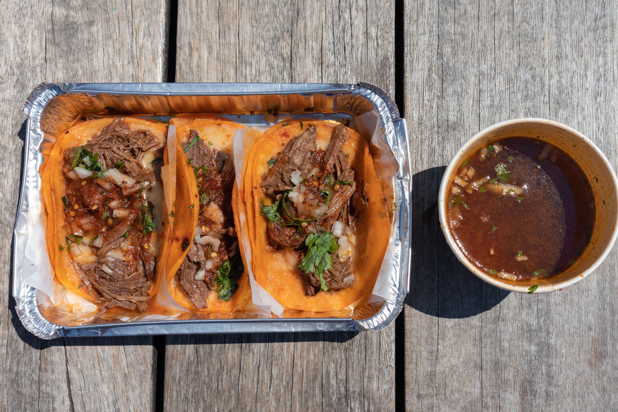 An overhead view of three beef birria tacos in an aluminum takeout container with a consomme soup on the side