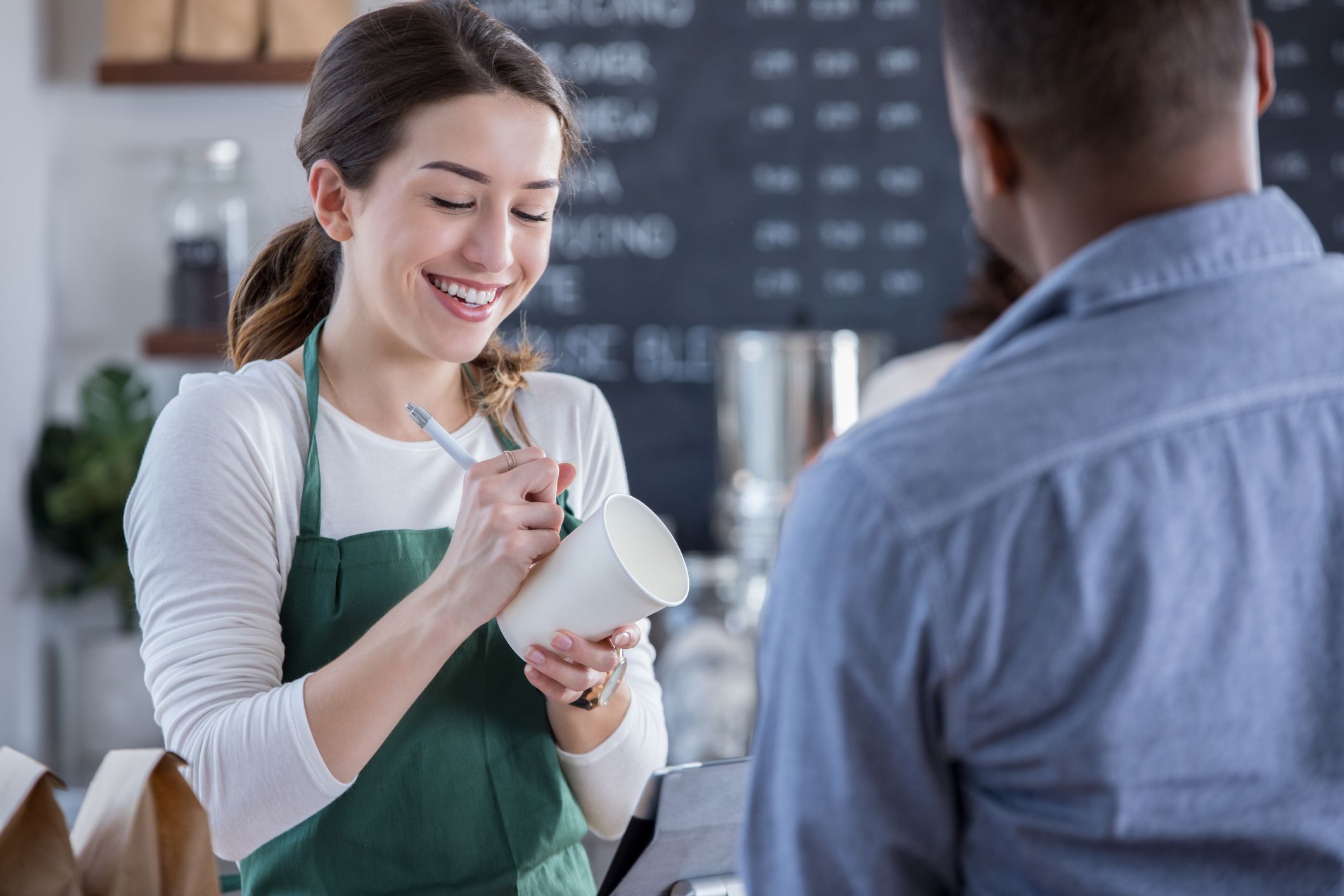 A smiling female coffee shop barista stands behind the checkout counter across from an unrecognizable patron. She looks down as she writes his order on a coffee cup.