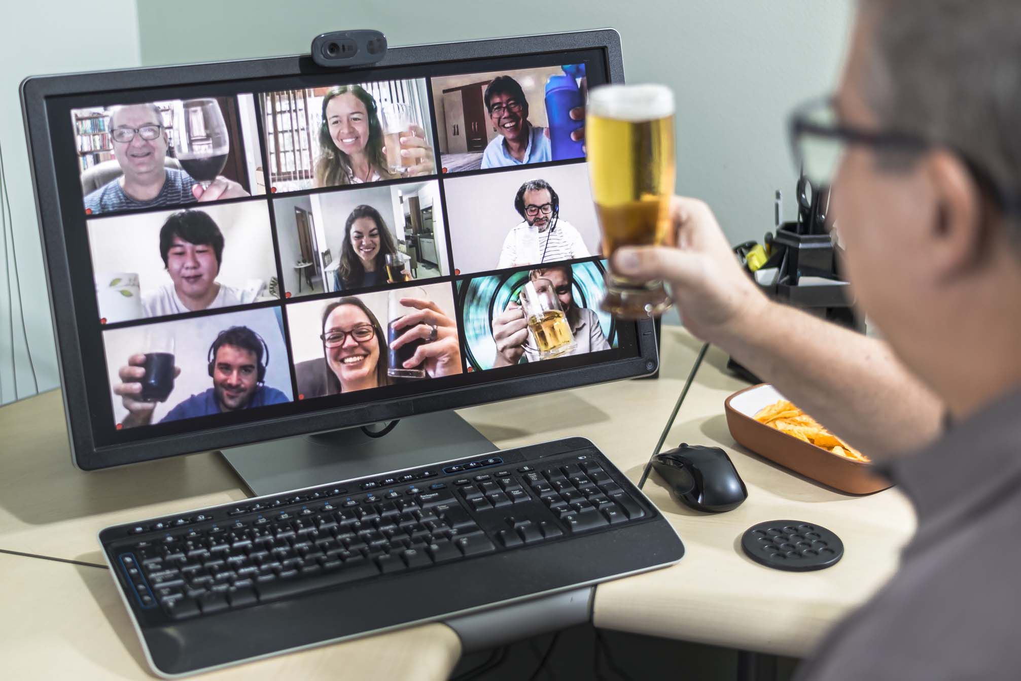 Computer screen of nine squares showing nine mixed people enjoying happy hour cocktails and beer with male watching screen on the right holding a beer for a toast, computer is on desk with food and work items like mouse and keyboard, during COVID-19