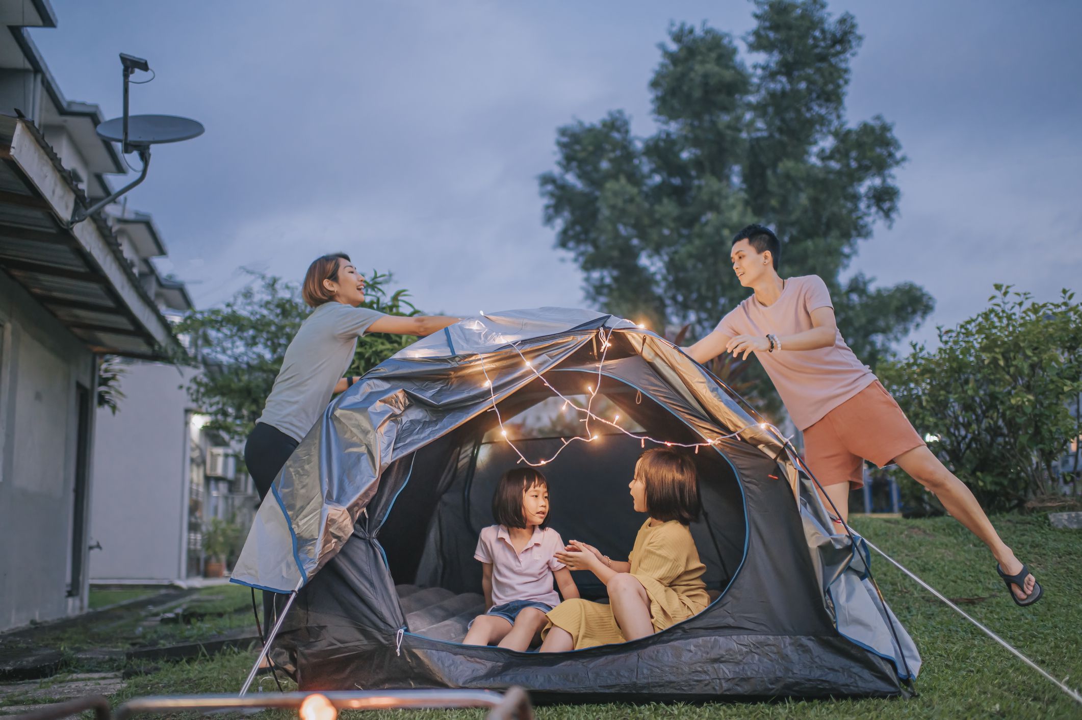 Couple putting on string light decorating tent, with two female children inside, at backyard of their house COVID-19 staycation, during the evening