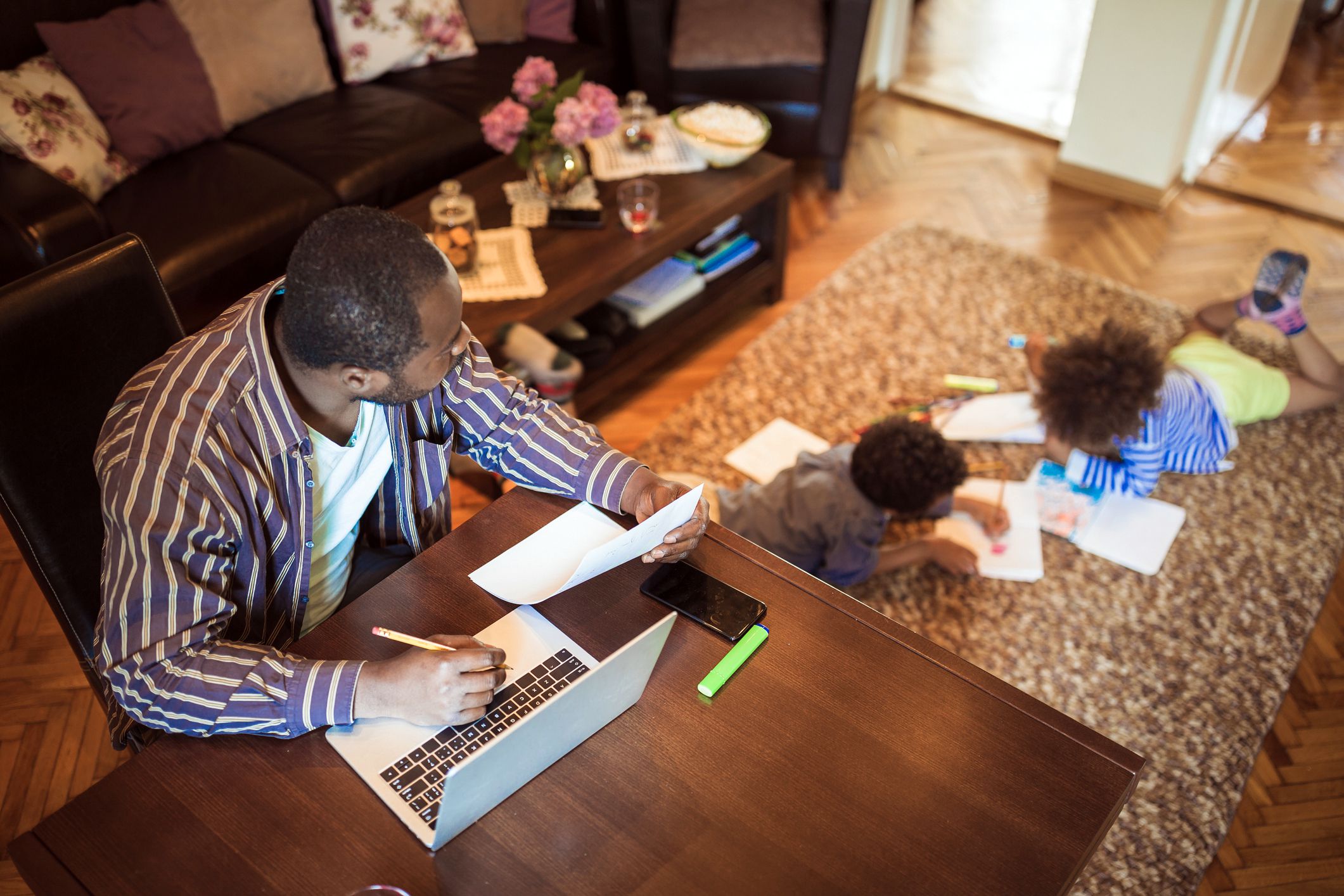 Father working from home on laptop on table while his two children are playing on the carpet in the living room