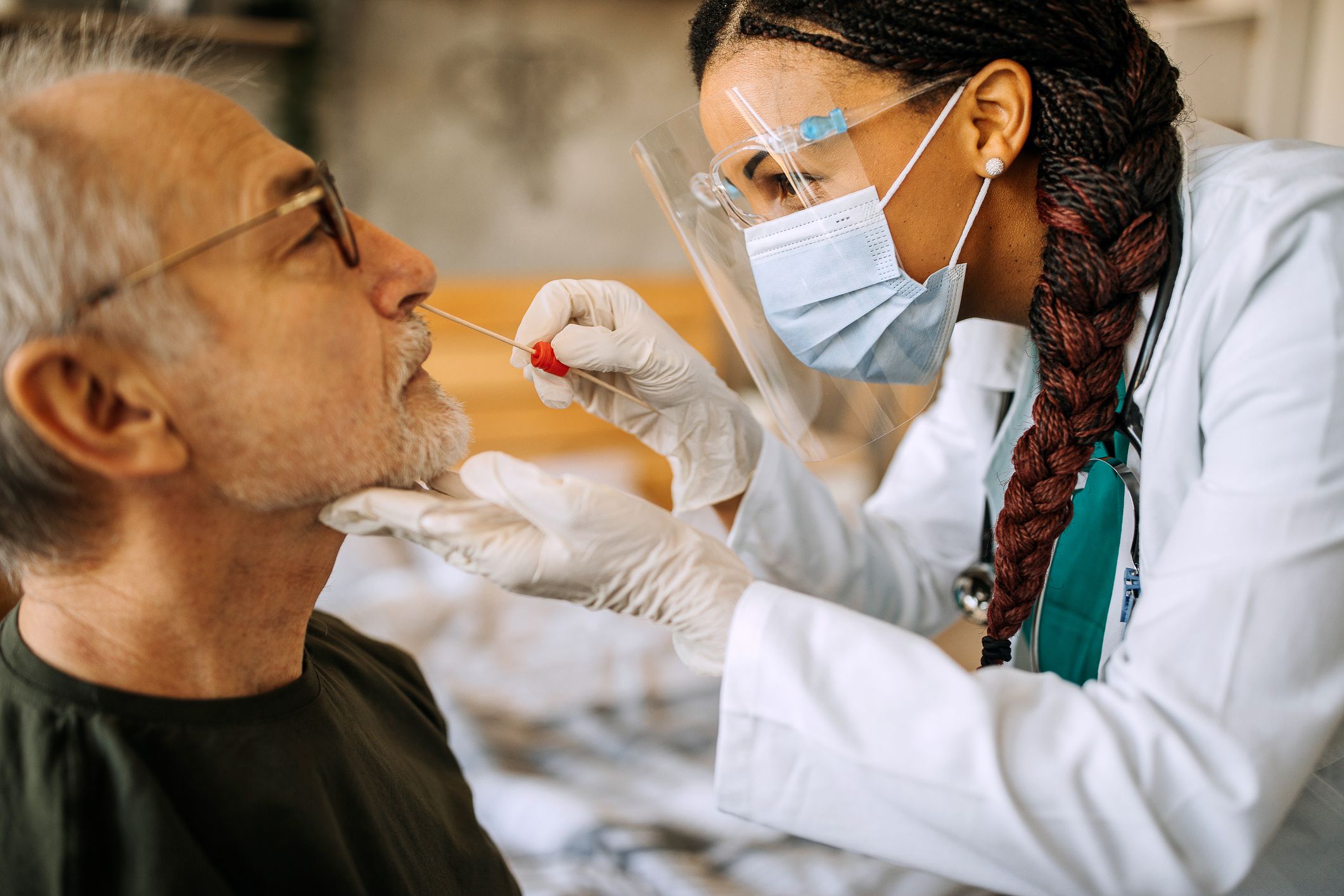 Female doctor is doing a home visit on senior man, taking a muscus sample from nose with swab for COVID-19 testing, with blurred background