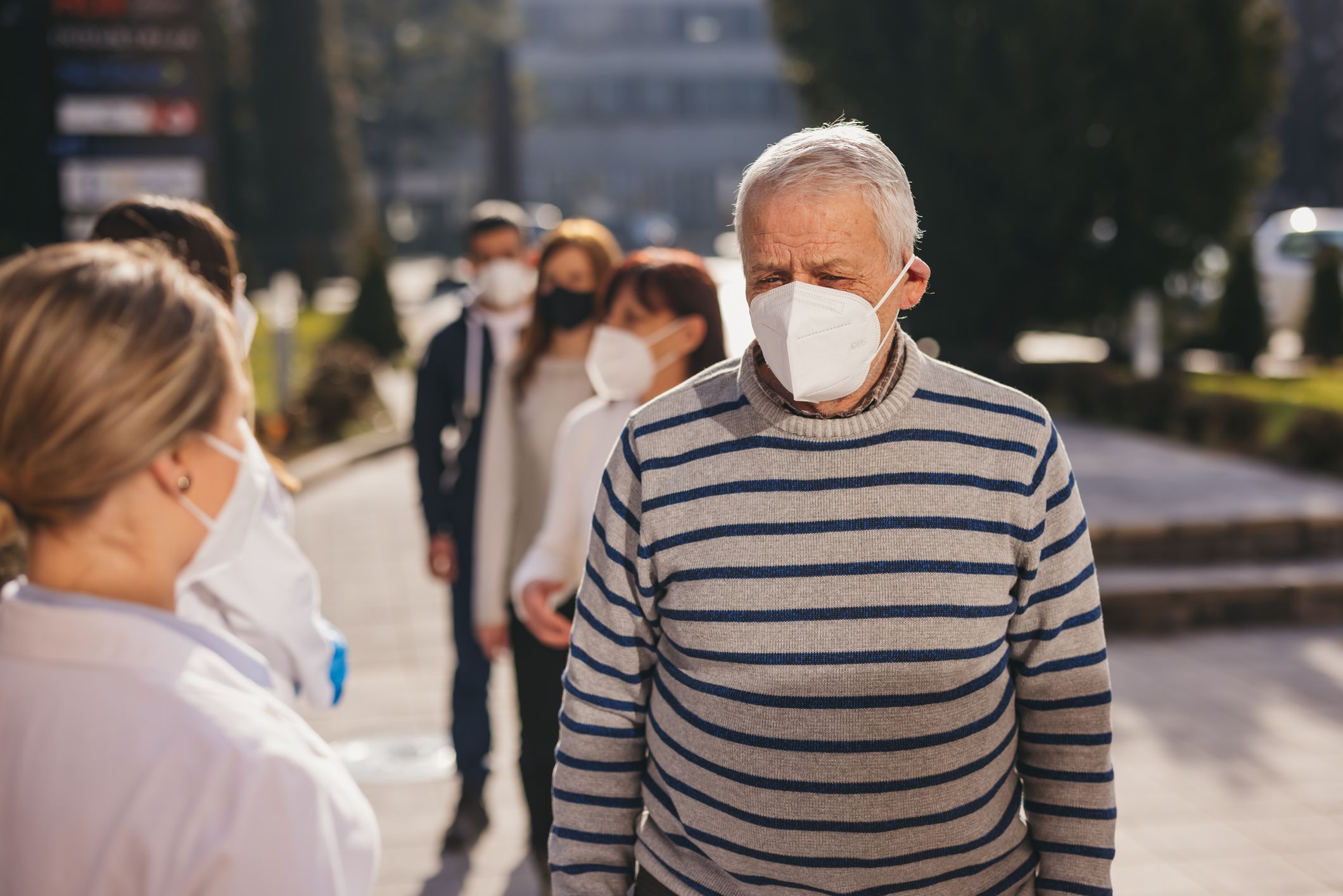 Group of mixed adult people with protective face masks standing in a line in front of a hospital and waiting for COVID-19 vaccination, while two young female doctors giving them explanations and directions