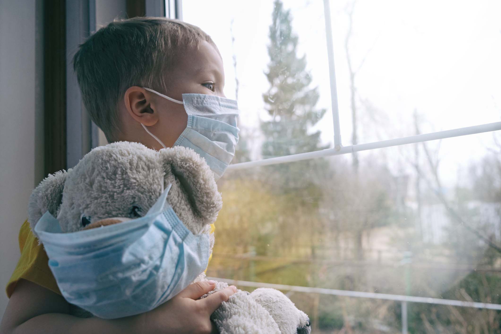 Hopeful male child and his teddy bear both in protective medical masks sits on windowsill and looks out window of late fall afternoon