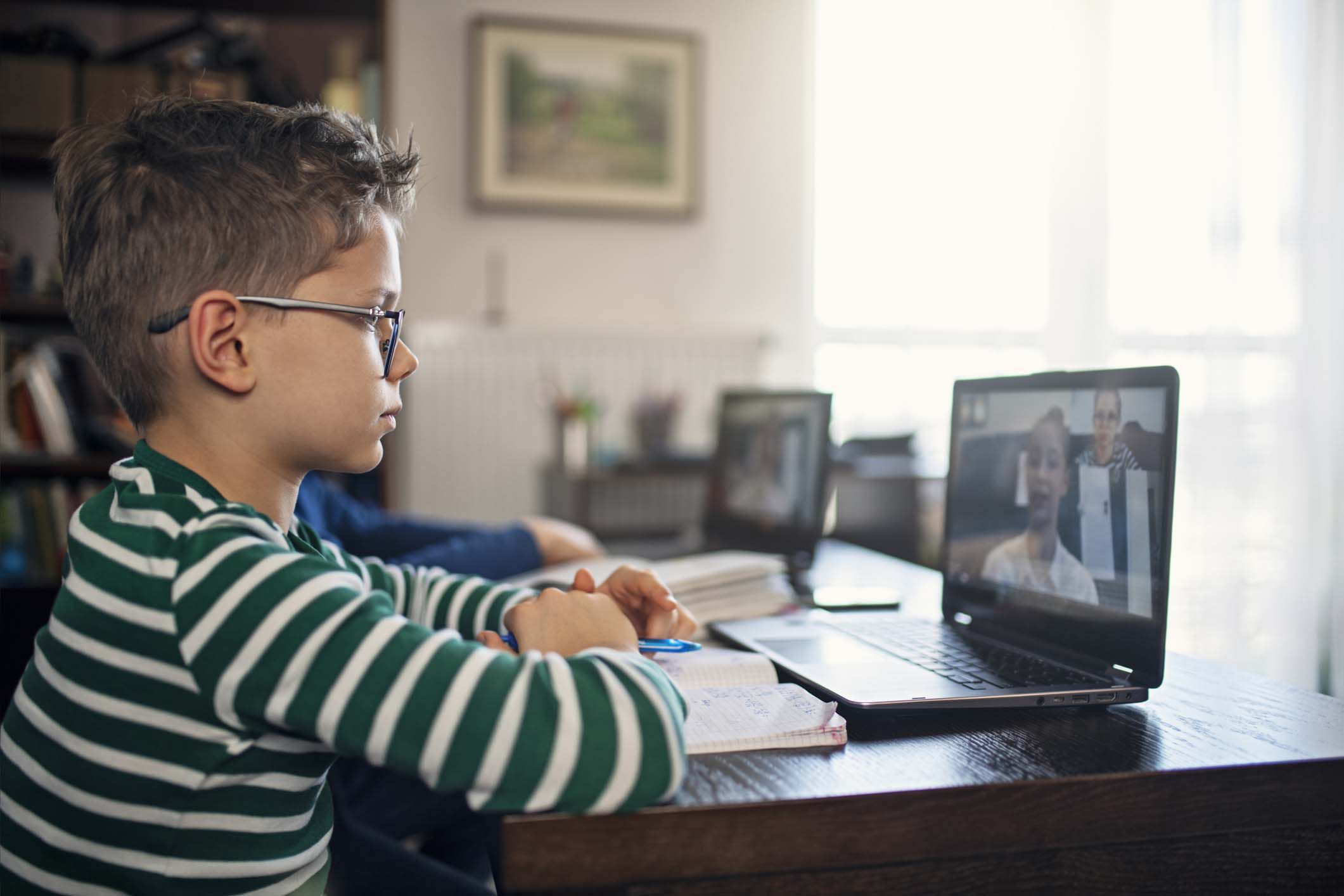 Male child at home attending online class on laptop on wooden table with light coming through big lights in the background