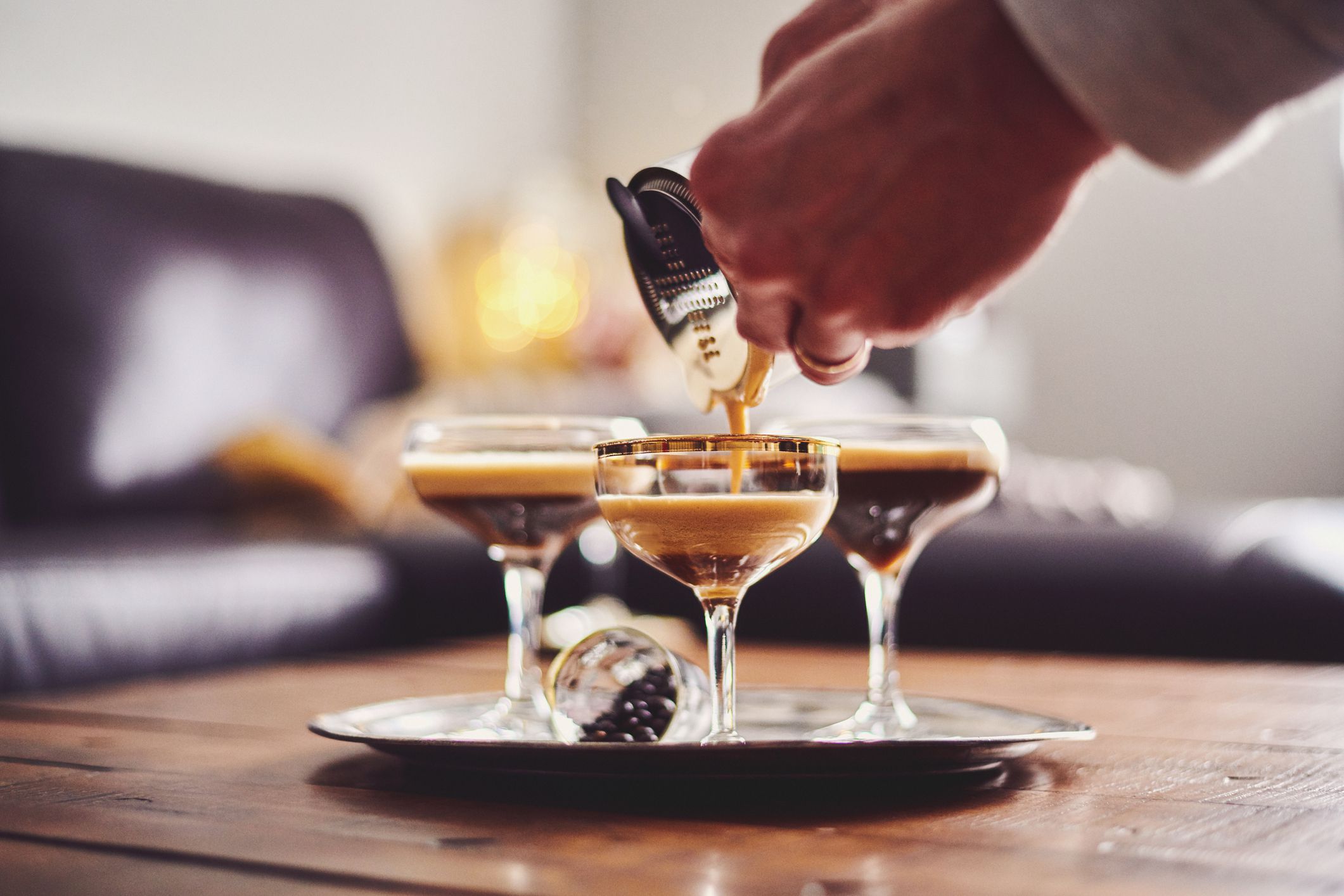 Male hands pouring espresso martini cocktail into glass, with two other glasses already filled, all on a tray on a wooden table in a living room, with a couch in the background
