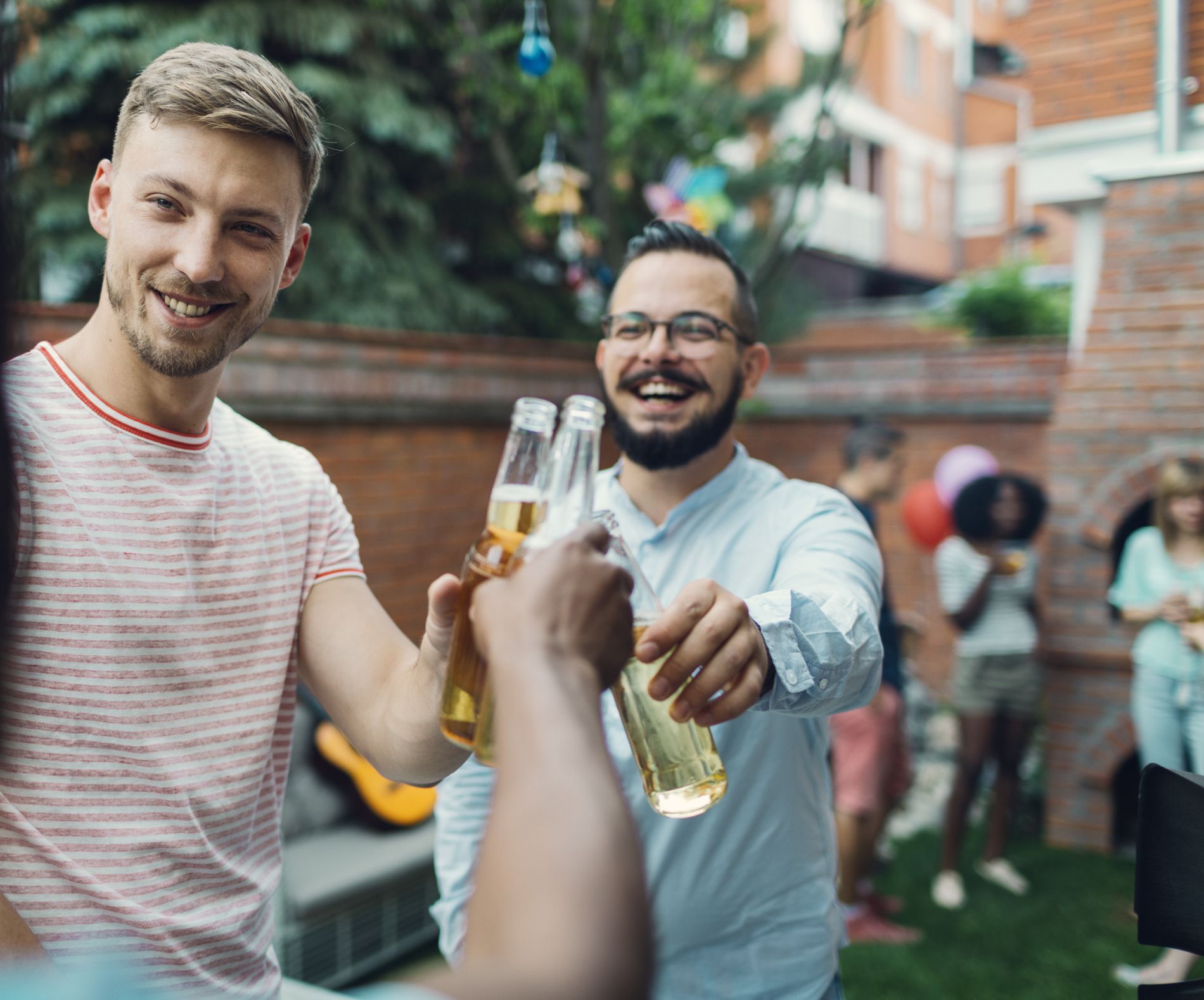 Three men toasting glass bottles of beer at COVID-19 barbecue party in brick walled backyard, one is only showing arm while other two are in close focus, friends talking in the background