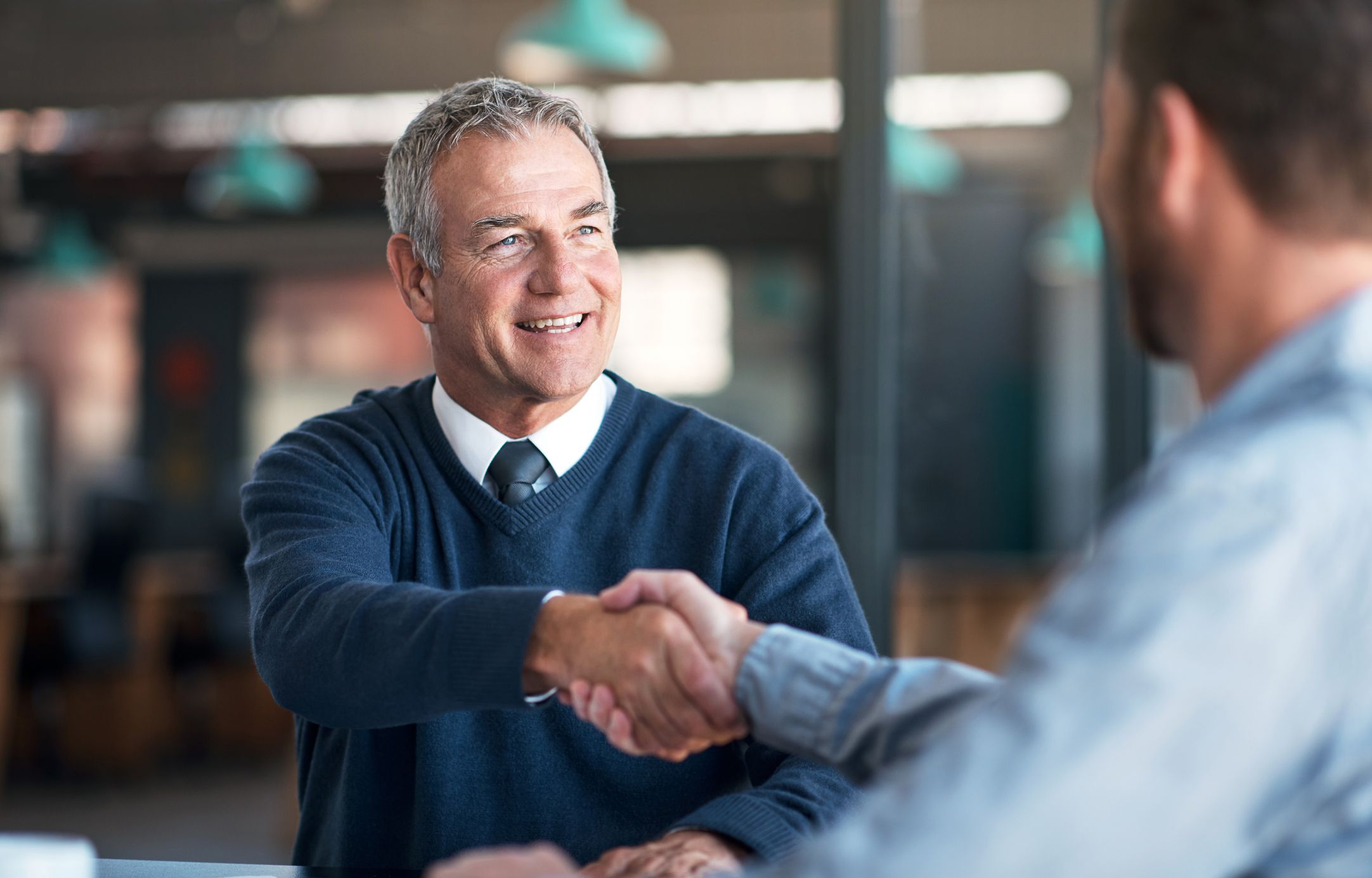 Two businessmen shaking hands in an office, showing one, who is smiling, and hand and arm of the other, blurred background