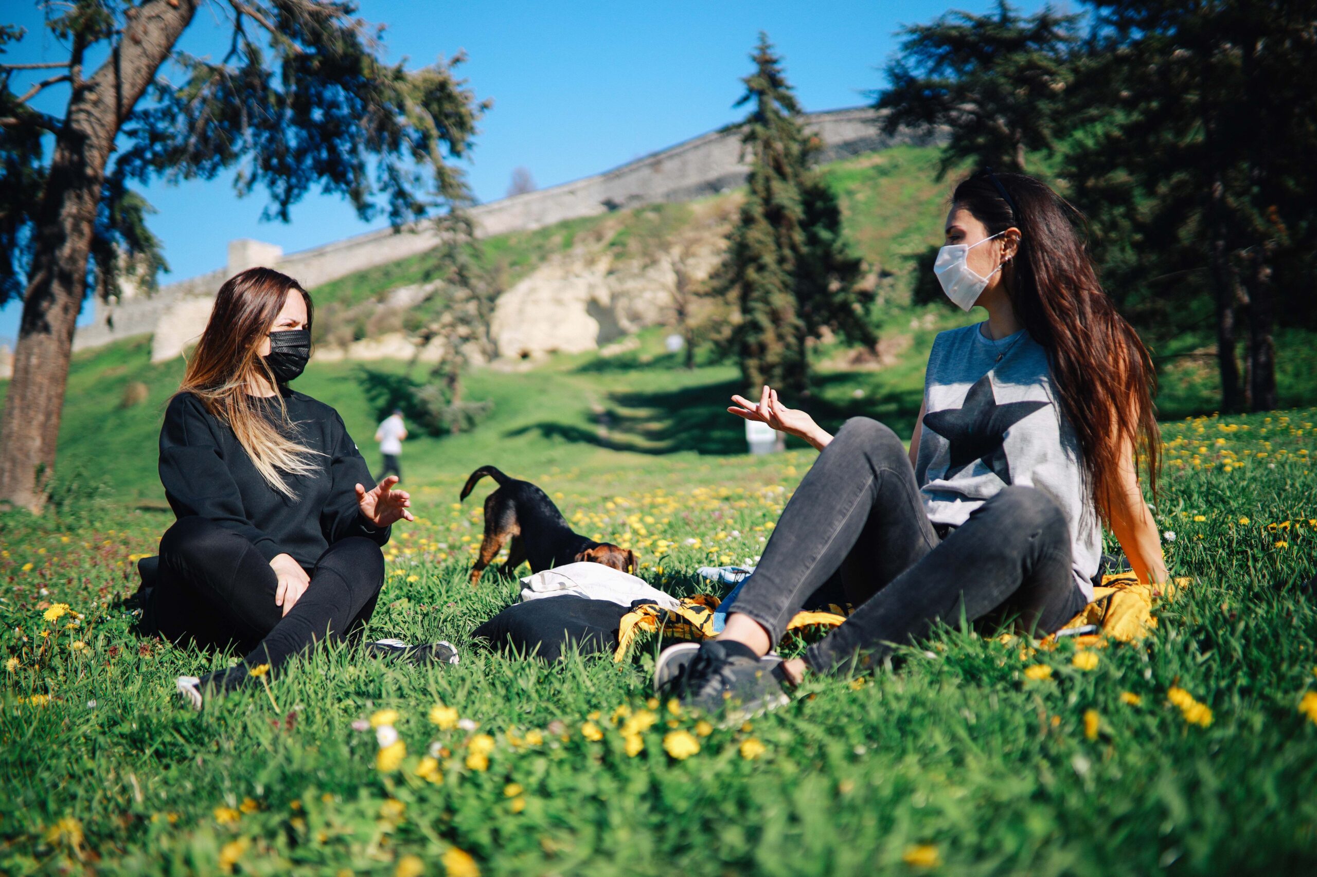 Two young women, both wearing protective masks, resting in the park with a dog duing COVID-19, hills and forest in the background
