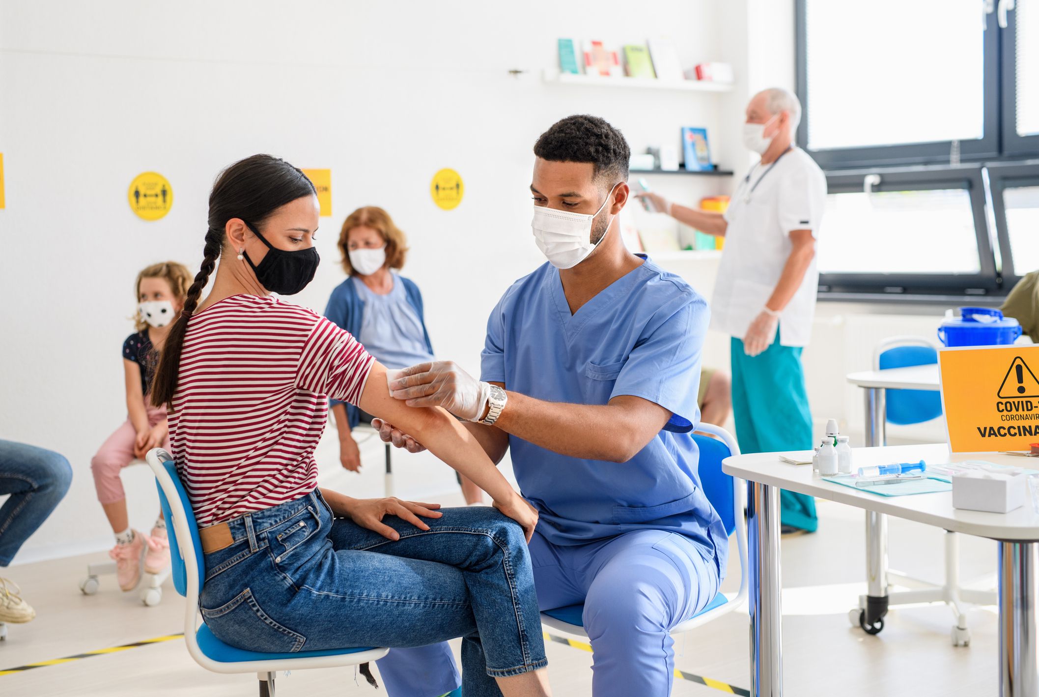 Young woman with face mask getting a COVID-19 vaccination from male nurse in a healthcare center with other patients getting vaccinations from nurses and doctors