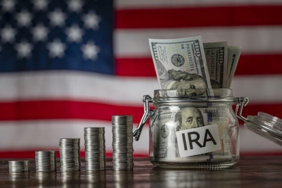 Five stacks of coins increasing in height next to a glass jar filled with hundred-dollar bills labeled “IRA,” with an American flag in the background.