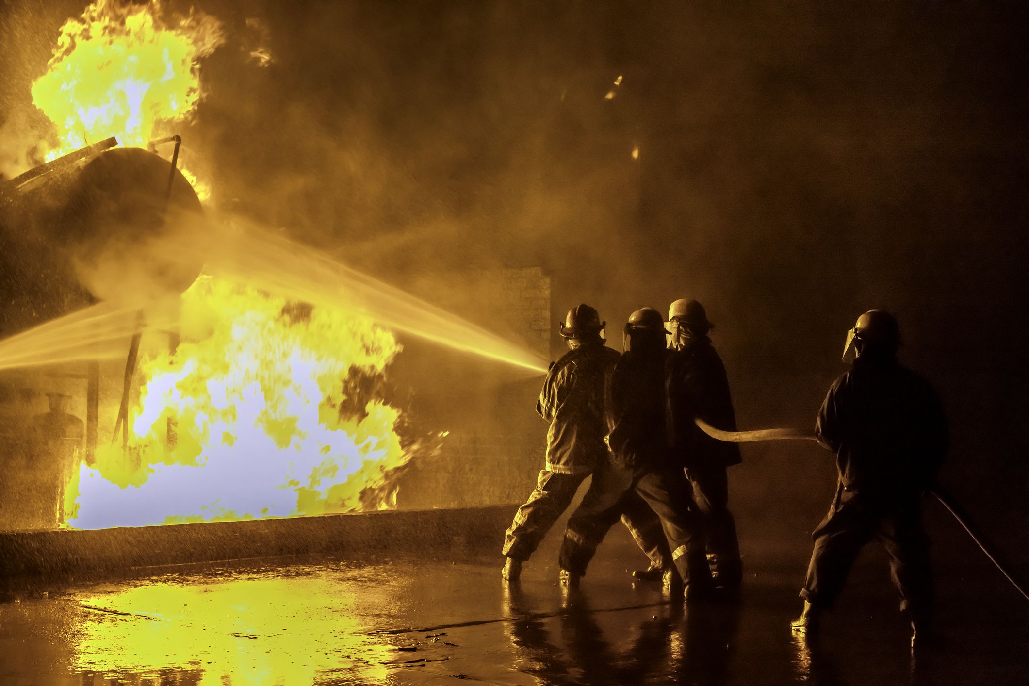 Firefighters practicing extinguishing an industrial fire at the fire station holding on to the hose spraying the fire at a storage tank.