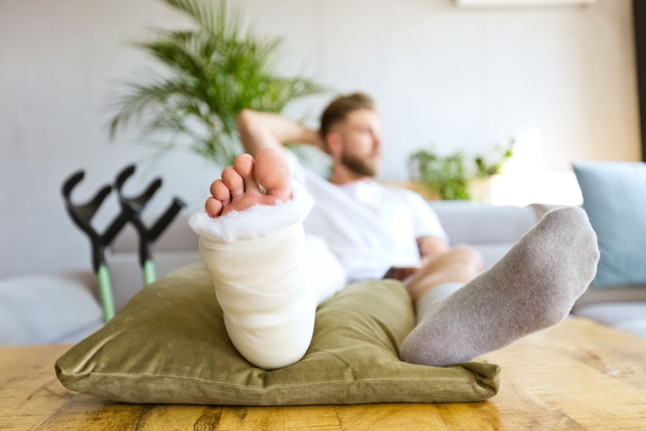 Young man with broken leg in plaster cast lying down on sofa at home, holding smart phone in hand and looking through window. Focus on leg.