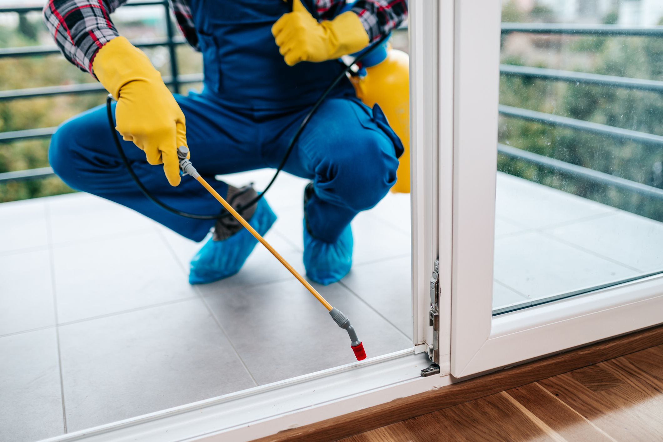 Professional exterminator in protective workwear spraying pesticide in apartment kitchen.