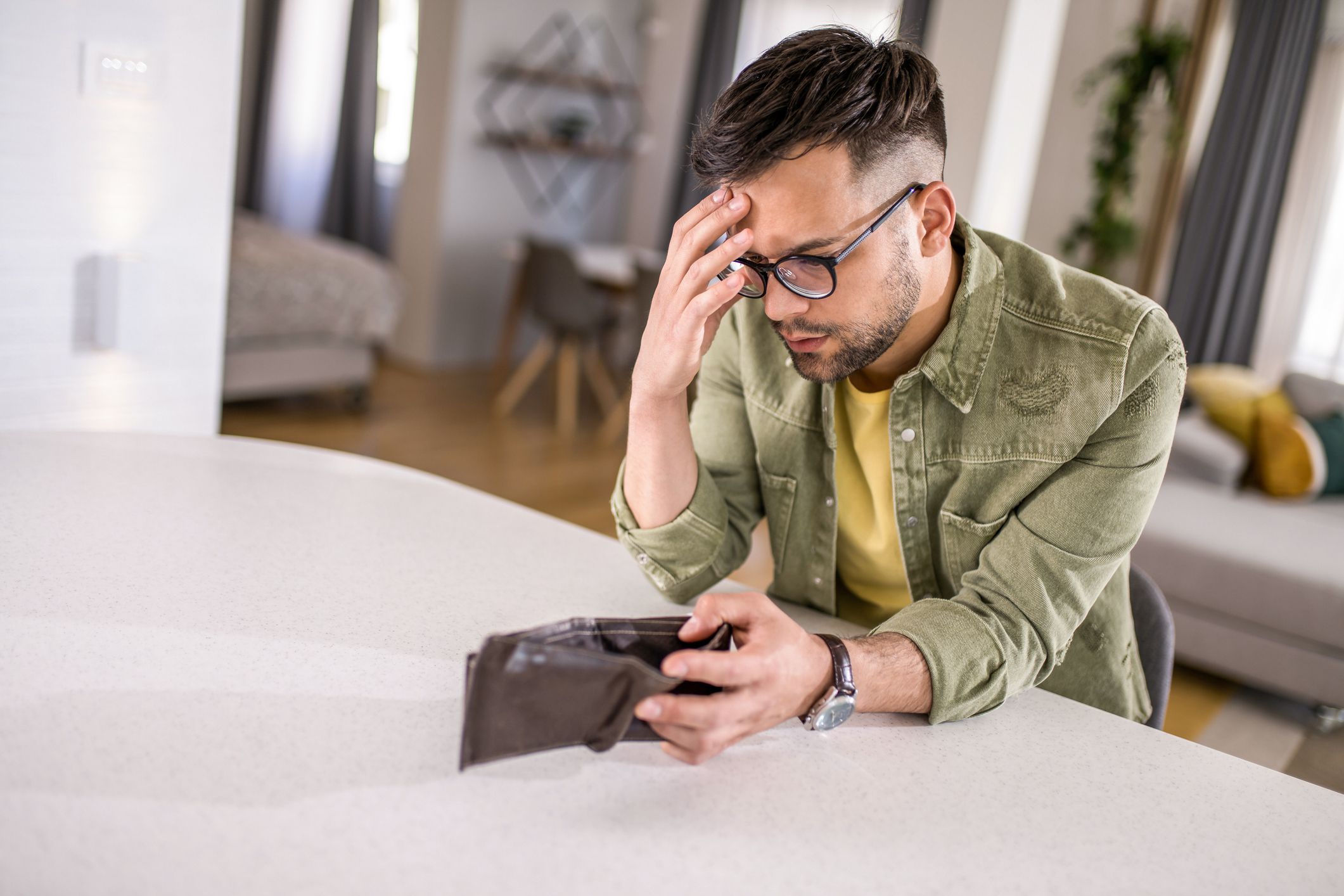 Young man sitting at home with an empty wallet. He is left without money and thinking what to do next.