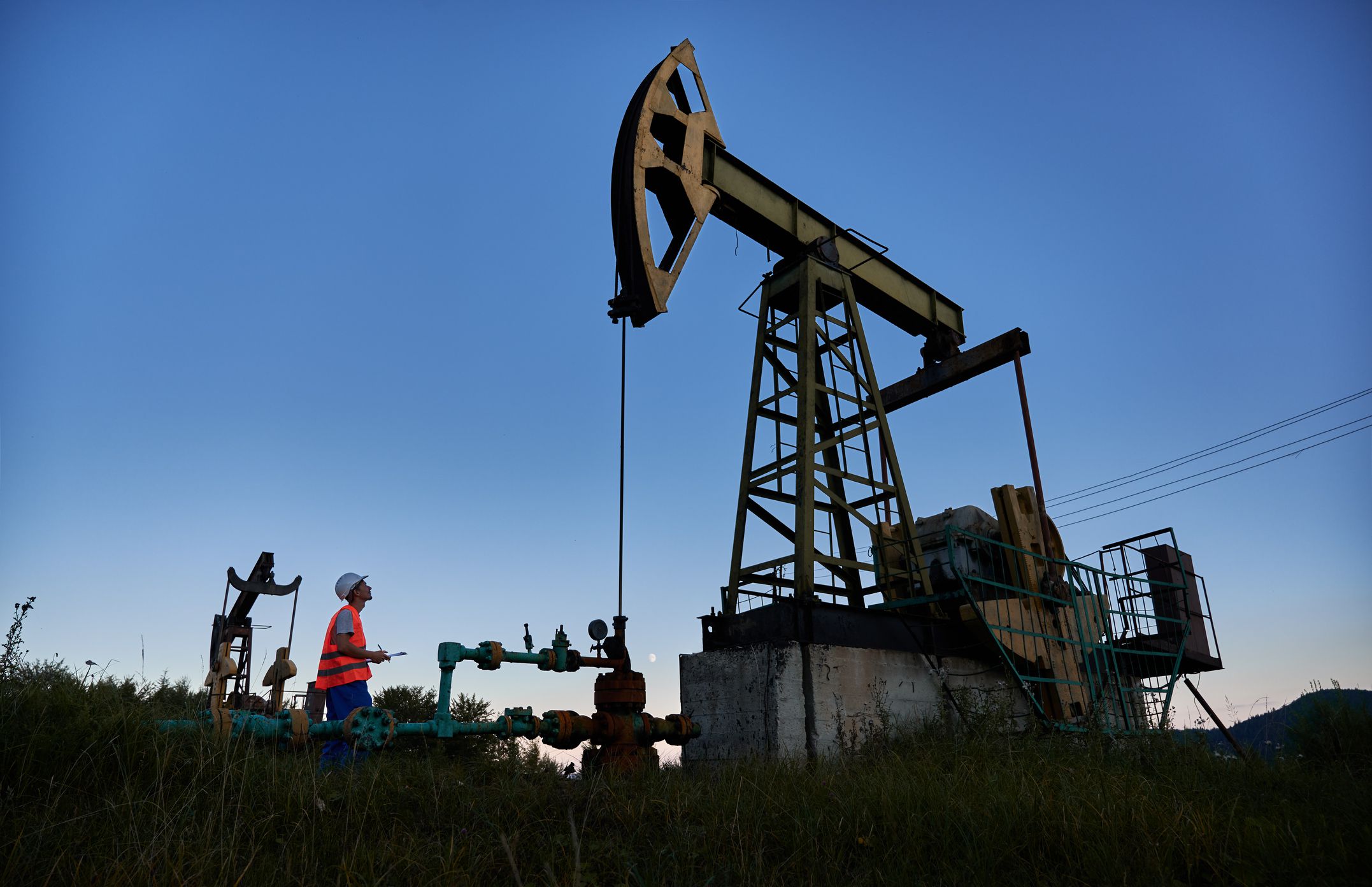 Male worker holding clipboard and looking at petroleum pump jack while working in oil field. Oil man standing near oil pumping unit under blue sky. Concept of oil extraction and petroleum industry.