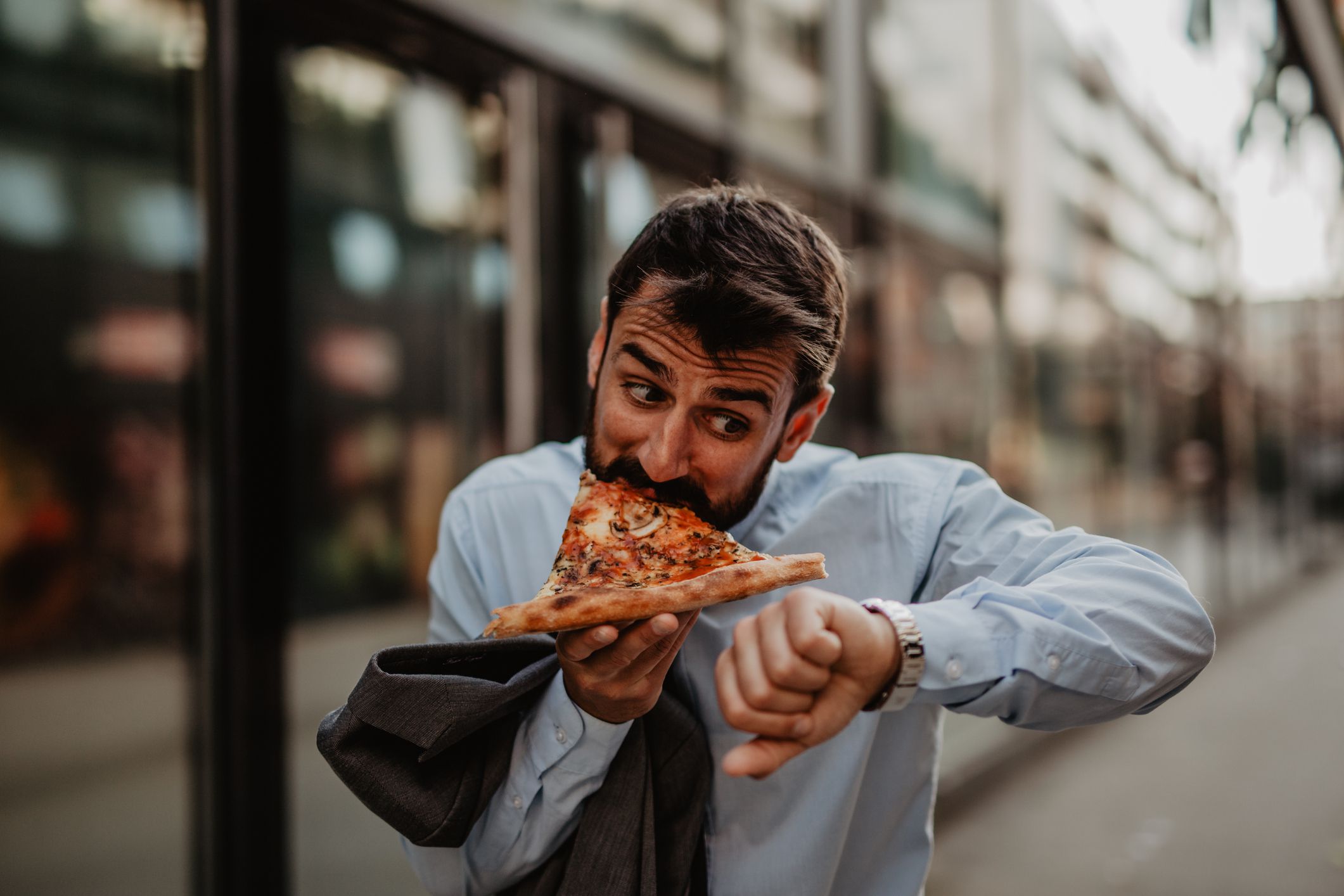 Image of a young businessman in rush, eating a pizza slice and checking the time on his wristwatch.