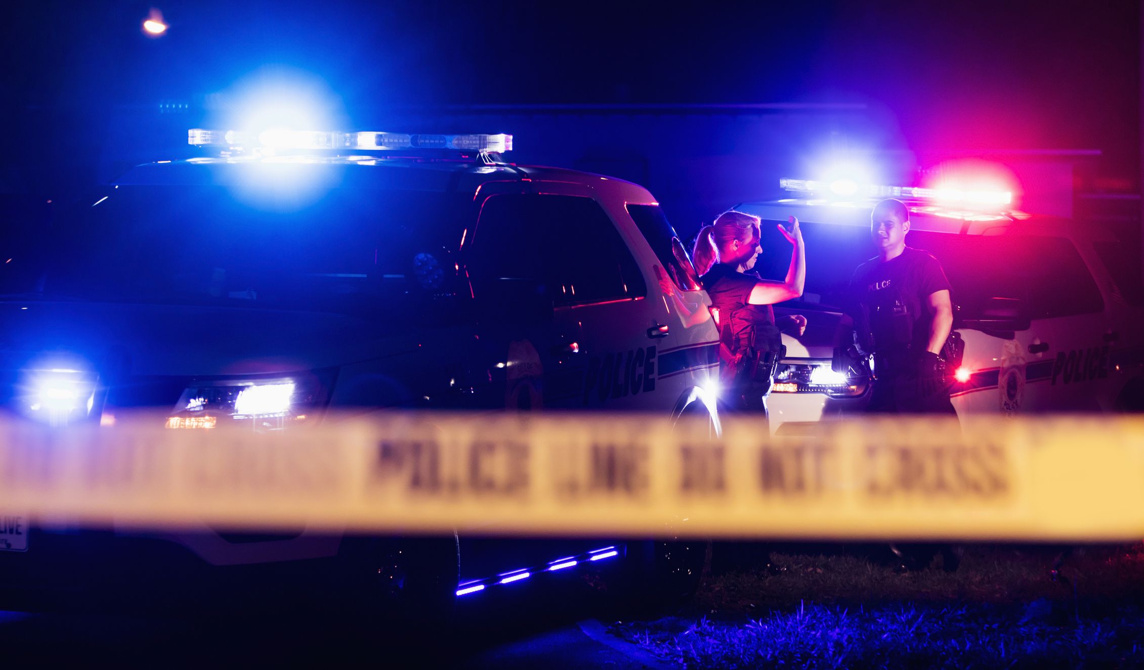 Two multi-ethnic police officers at night wearing bulletproof vests, standing beside police cars with emergency lights flashing, behind cordon tape. The policewoman is in her 40s and her Hispanic, male partner is in his 30s.