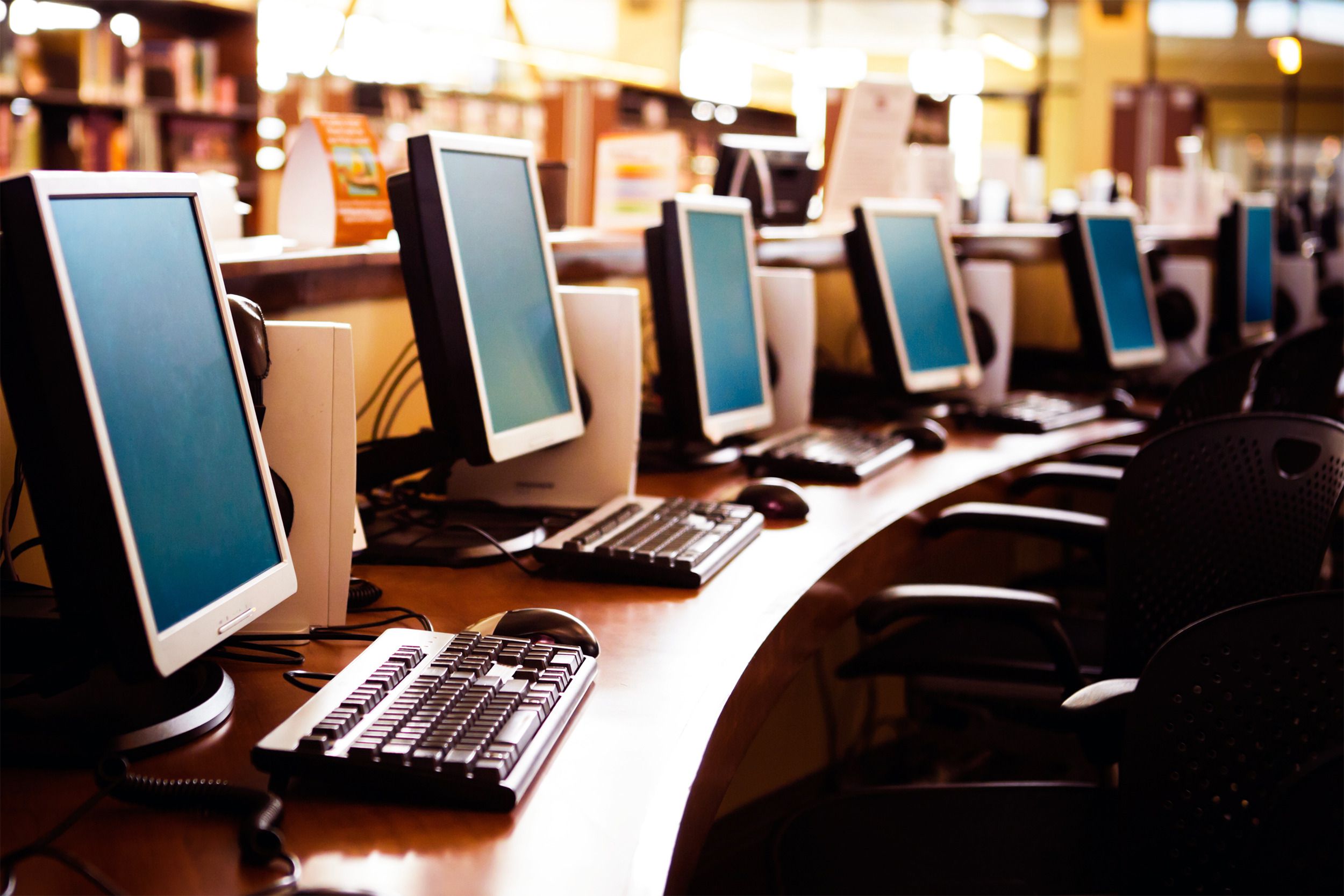 A Row of desktop computers with seats on a table in a public library with a blurred background of shelves of books