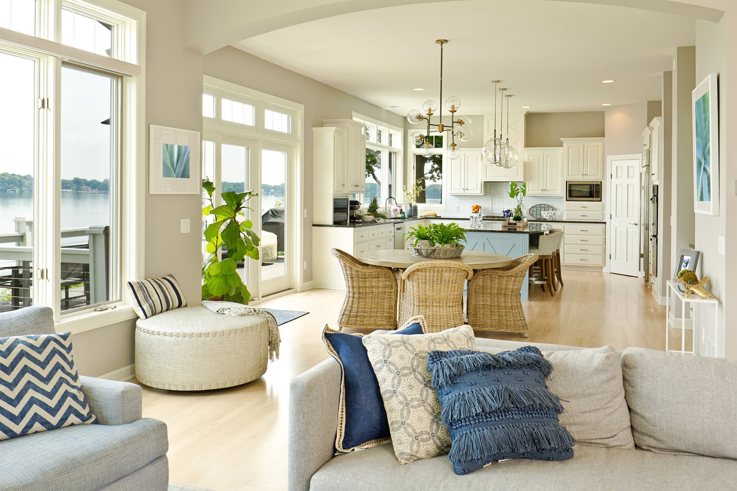 A staged kitchen and living room in a modern home in neutral light greys and beiges with navy blue accents, with the patio and a lake outside in the background
