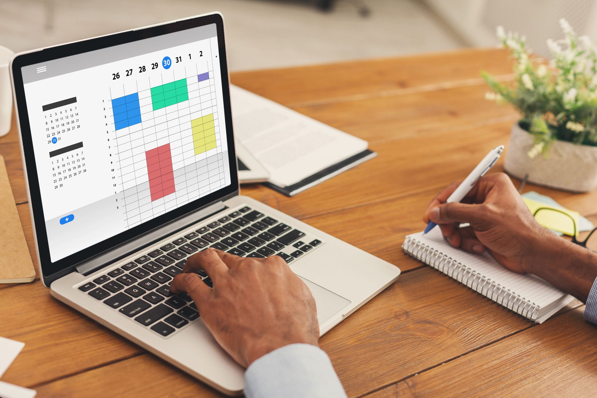 Businessman using online planner on laptop while writing notes with papers and notebooks on desk with a blurred background