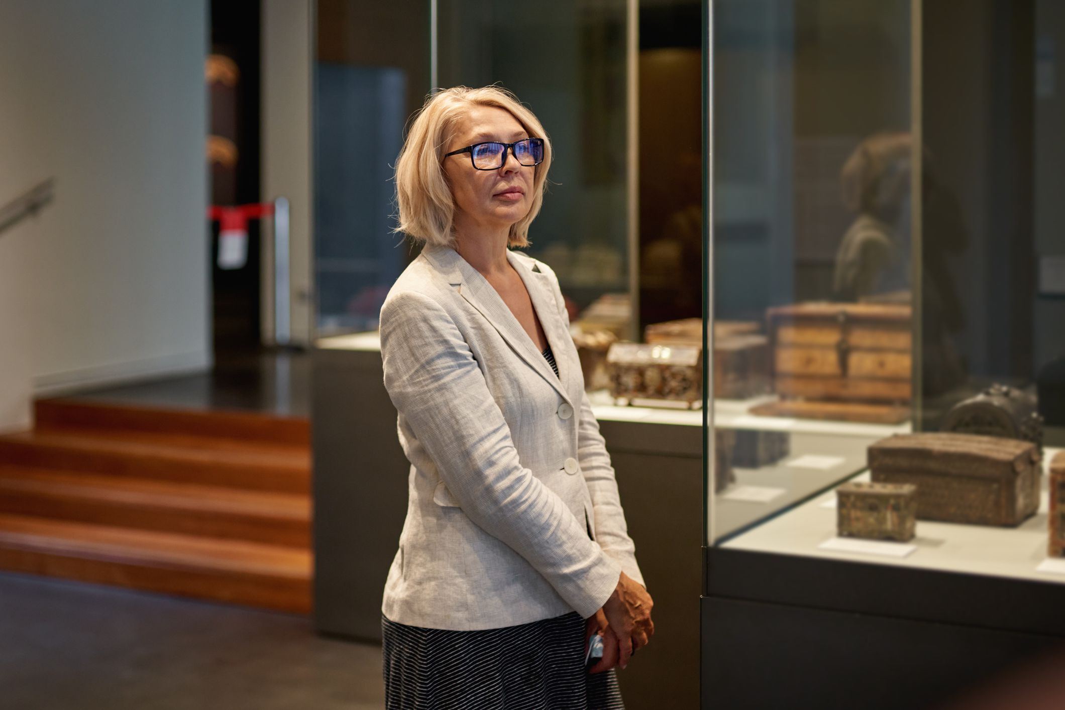 Classy mature woman exploring medieval exhibits in a museum through glass cases with stairs and another room in the background