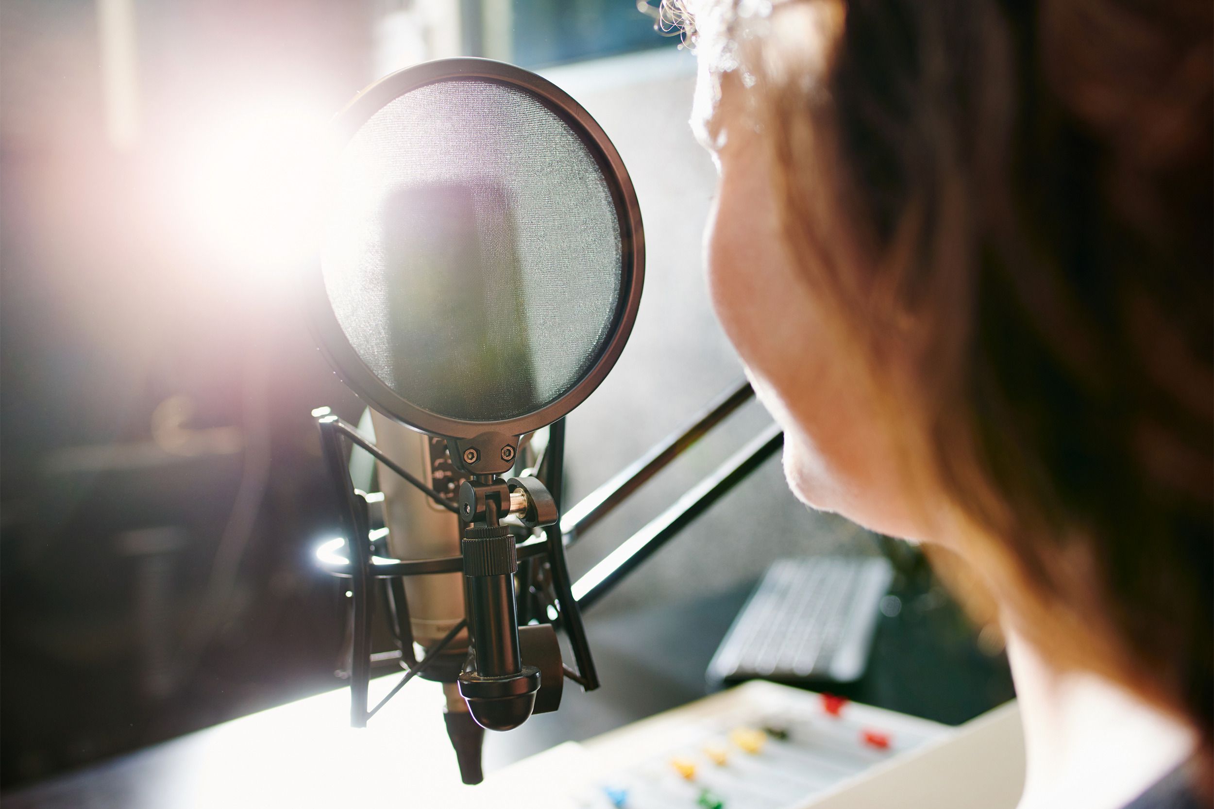 Closeup of a microphone while a mature woman speaking into it in a studio with a bright light with a blurred background of audio equipment