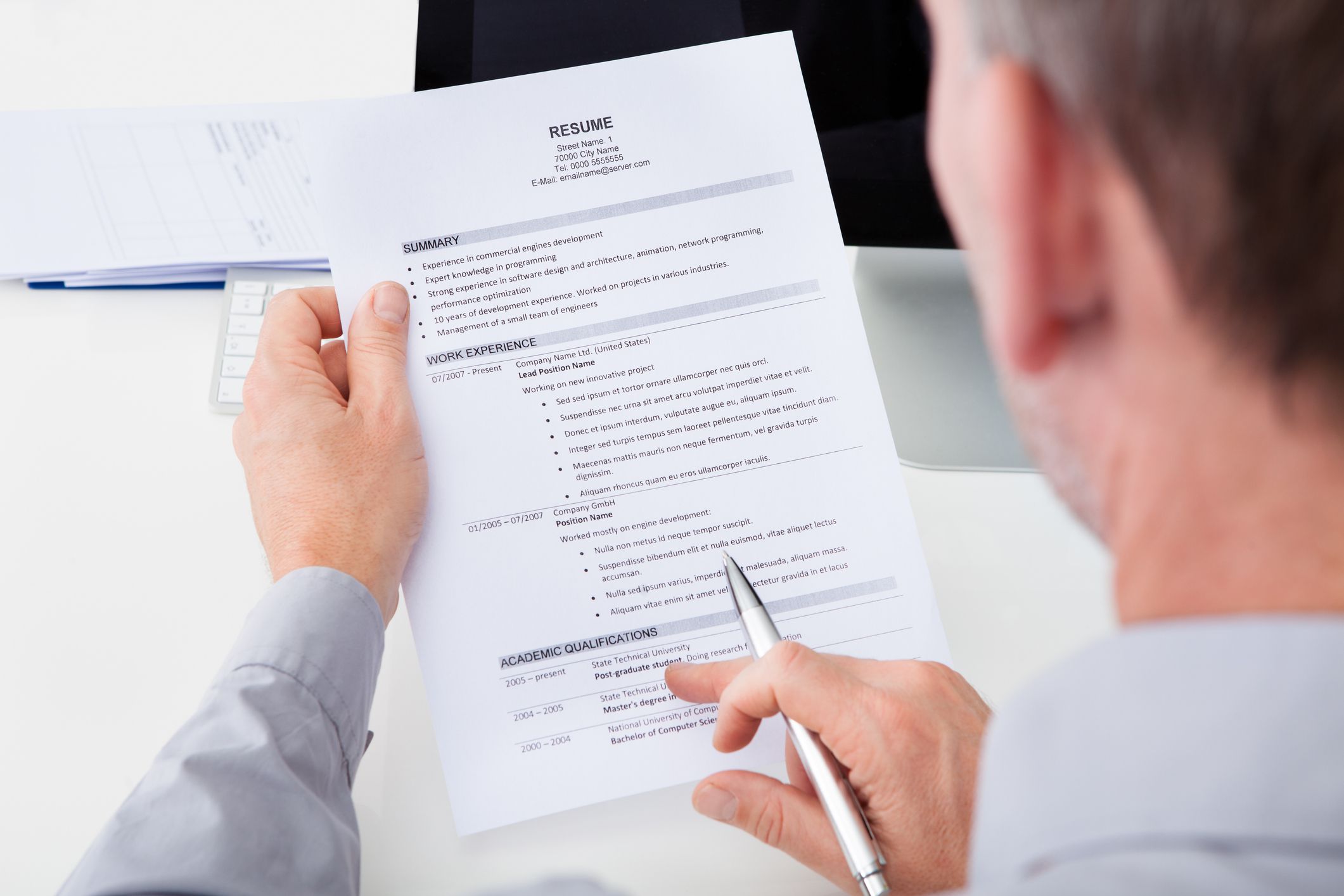 Closeup of a senior male recruiter looking at a resume with one hand holding it, while the other hand is holding a pen with a desktop computer, papers and a white table