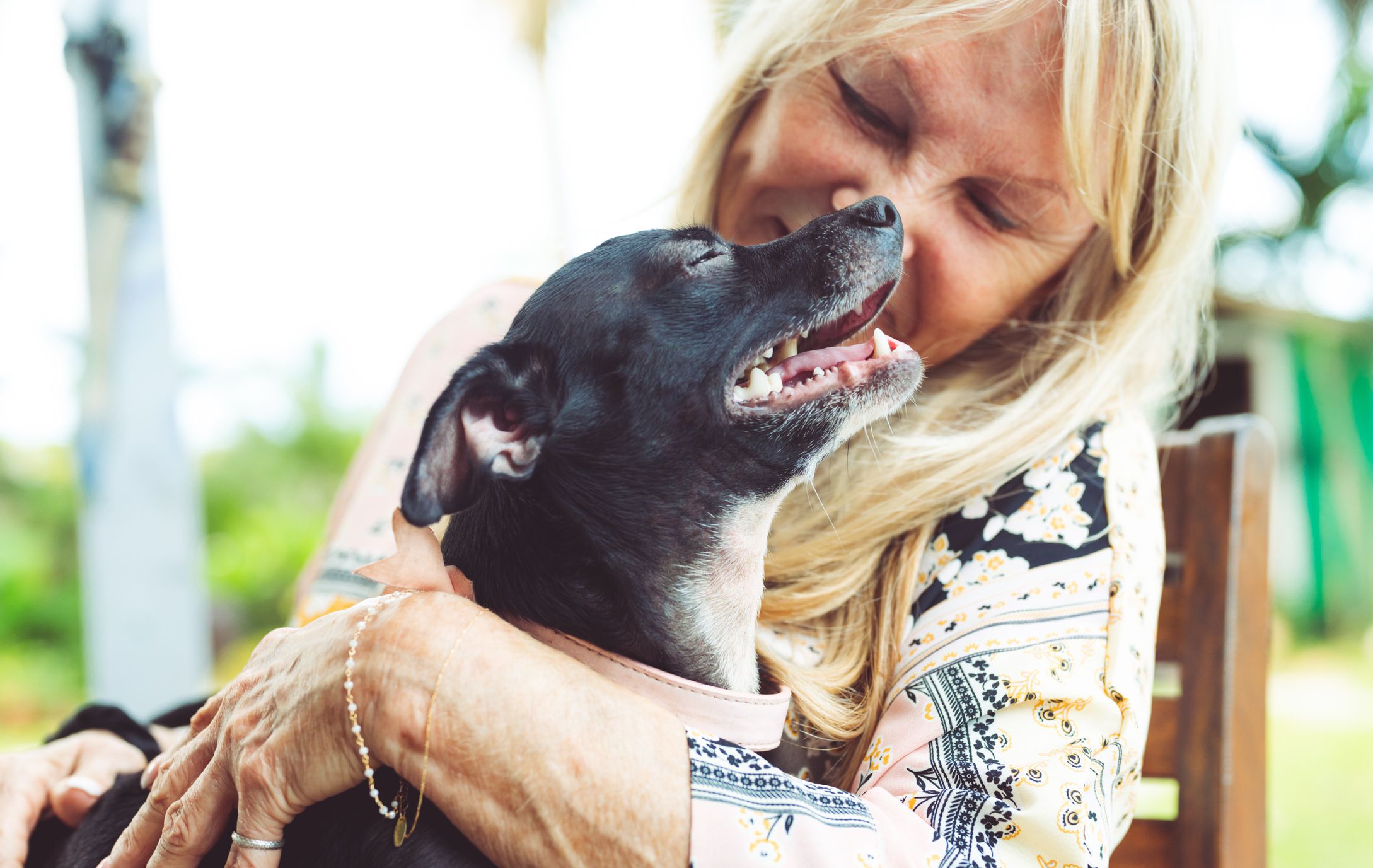 Closeup of a senior woman hugging her black dog while sitting in a chair outside with a blurred background of trees and plants