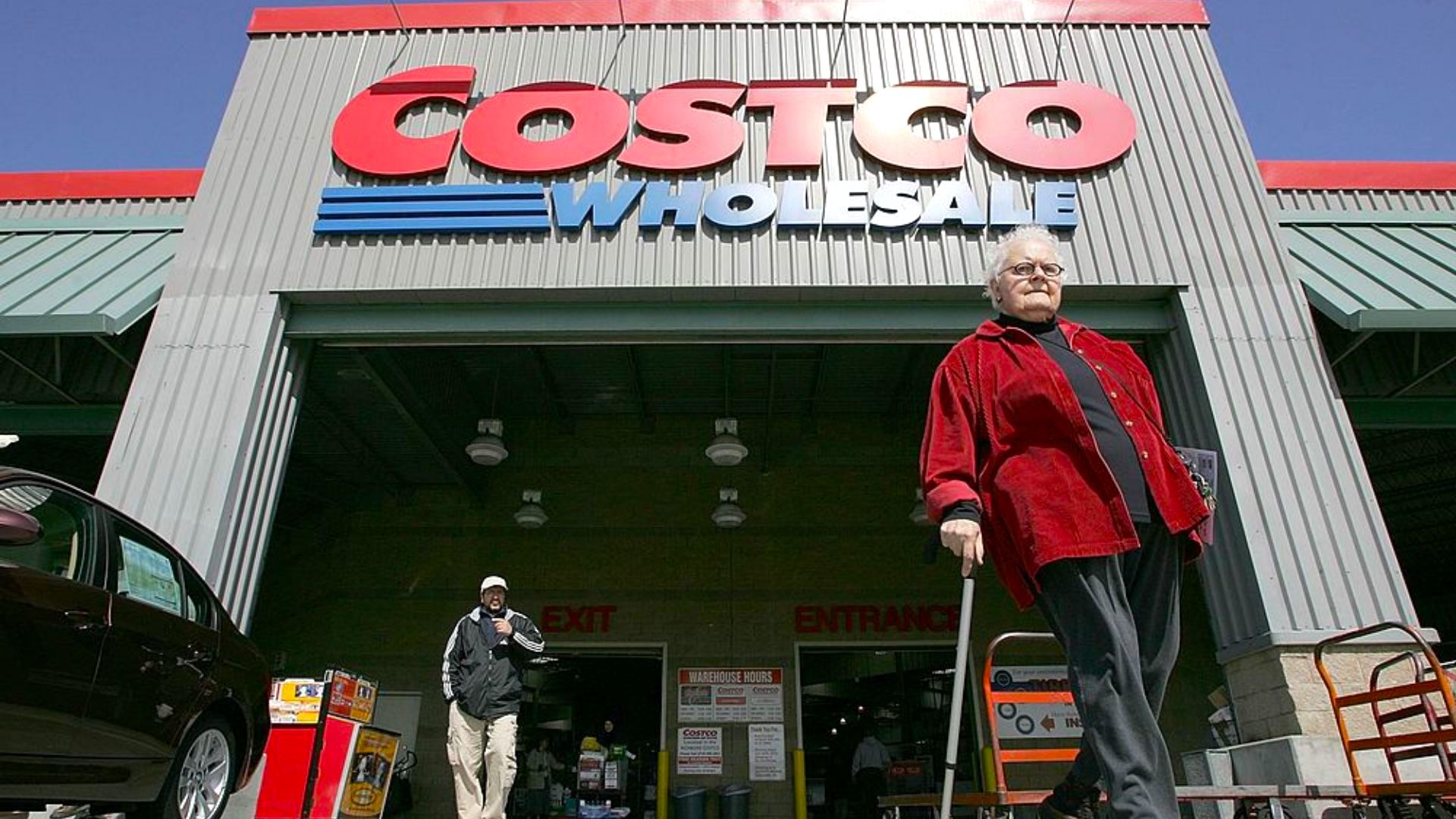 An older woman in a red jacket walks with a cane outside a Costco Wholesale store, possibly on her way to check out exclusive Costco deals for seniors. Another person and shopping carts are visible under the large Costco sign.