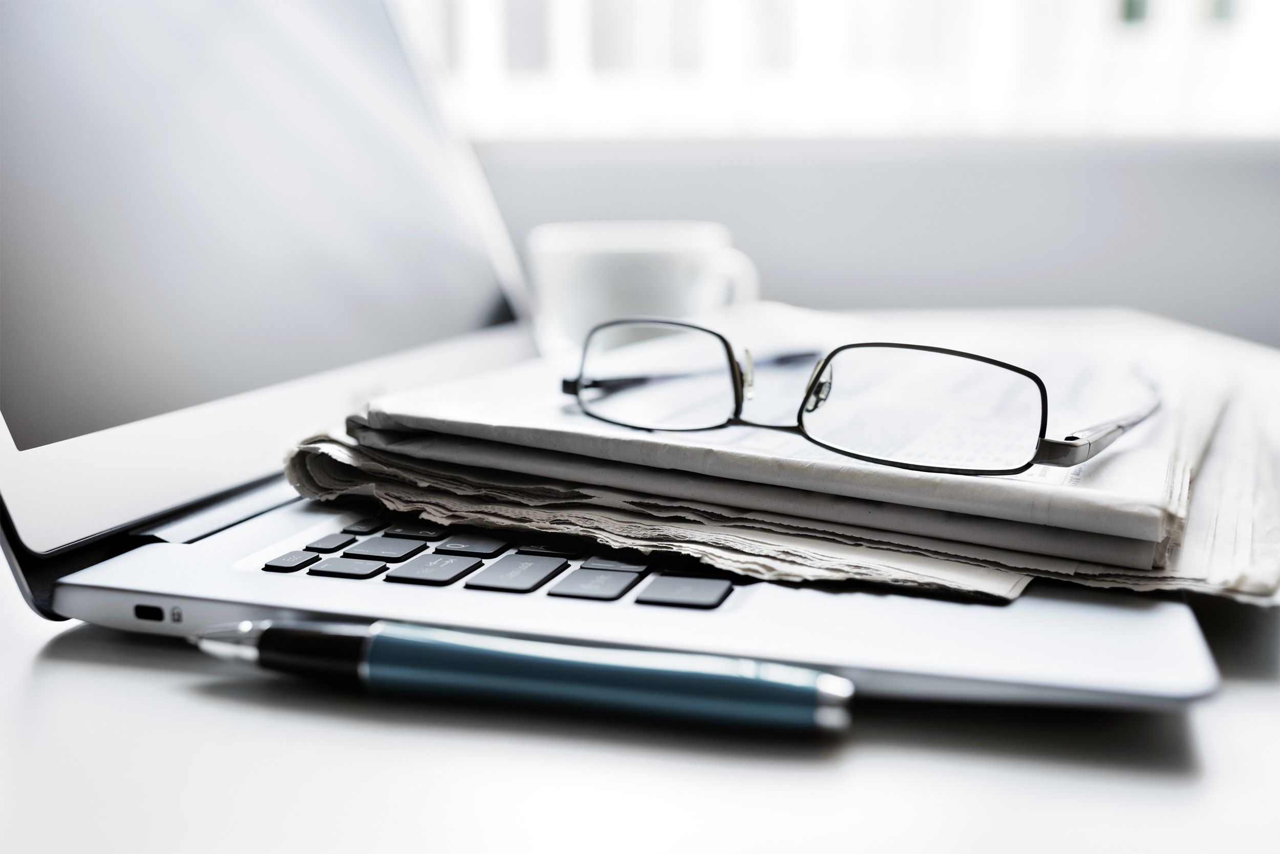 Glasses on top of a stack of newpapers on a laptop on a table, grey-tones with a blurred background of a coffee cup and window