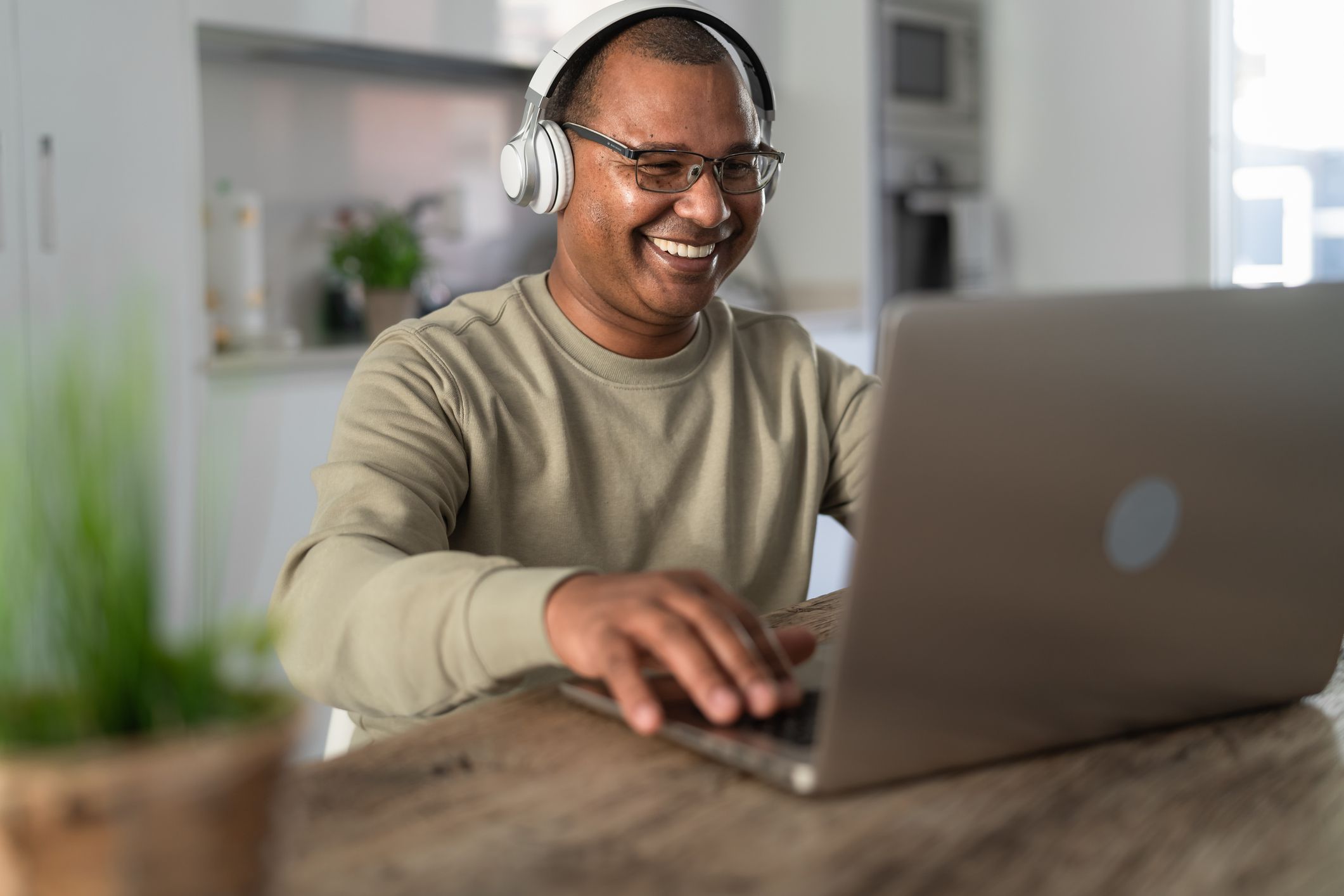 Happy senior man having fun using headphones and laptop at home on a wooden table with a blurred backgrounf od the kitchen