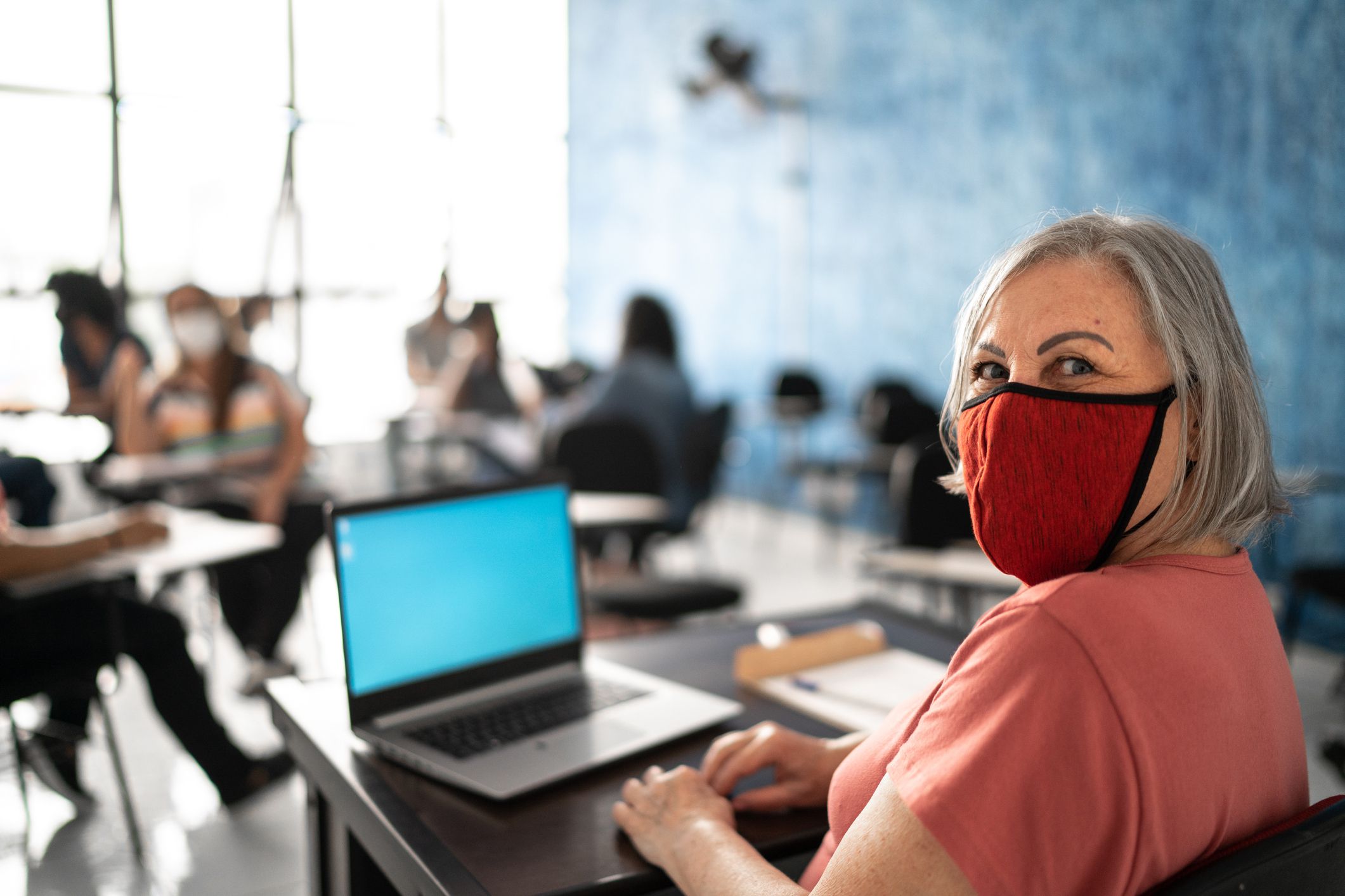 Mature female teacher with a face mask and laptop on a teacher desk looking at viewer with a blurred background of a classroom of children sitting at individual desks