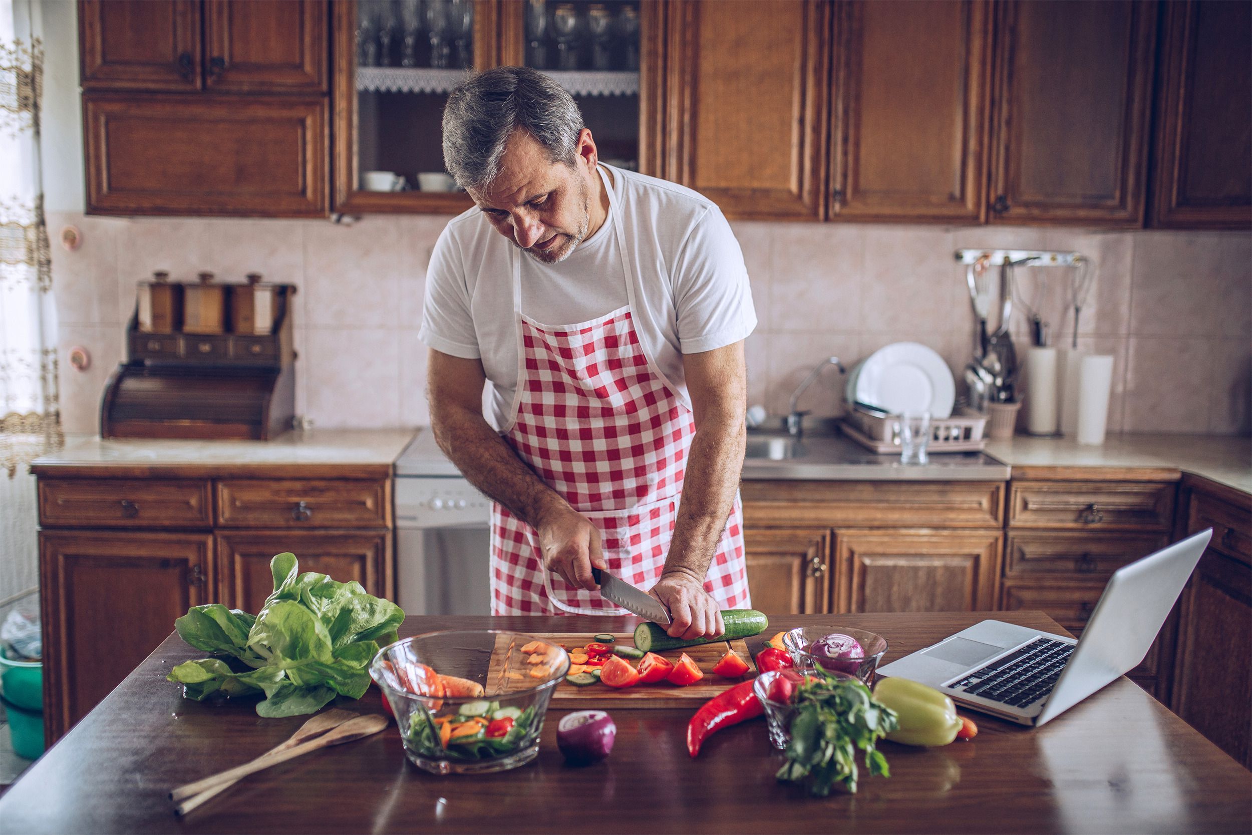 Mature male chef preparing numerous vegetables on a table with a laptop in a kitchen at a home with the kitchen counter is in the background