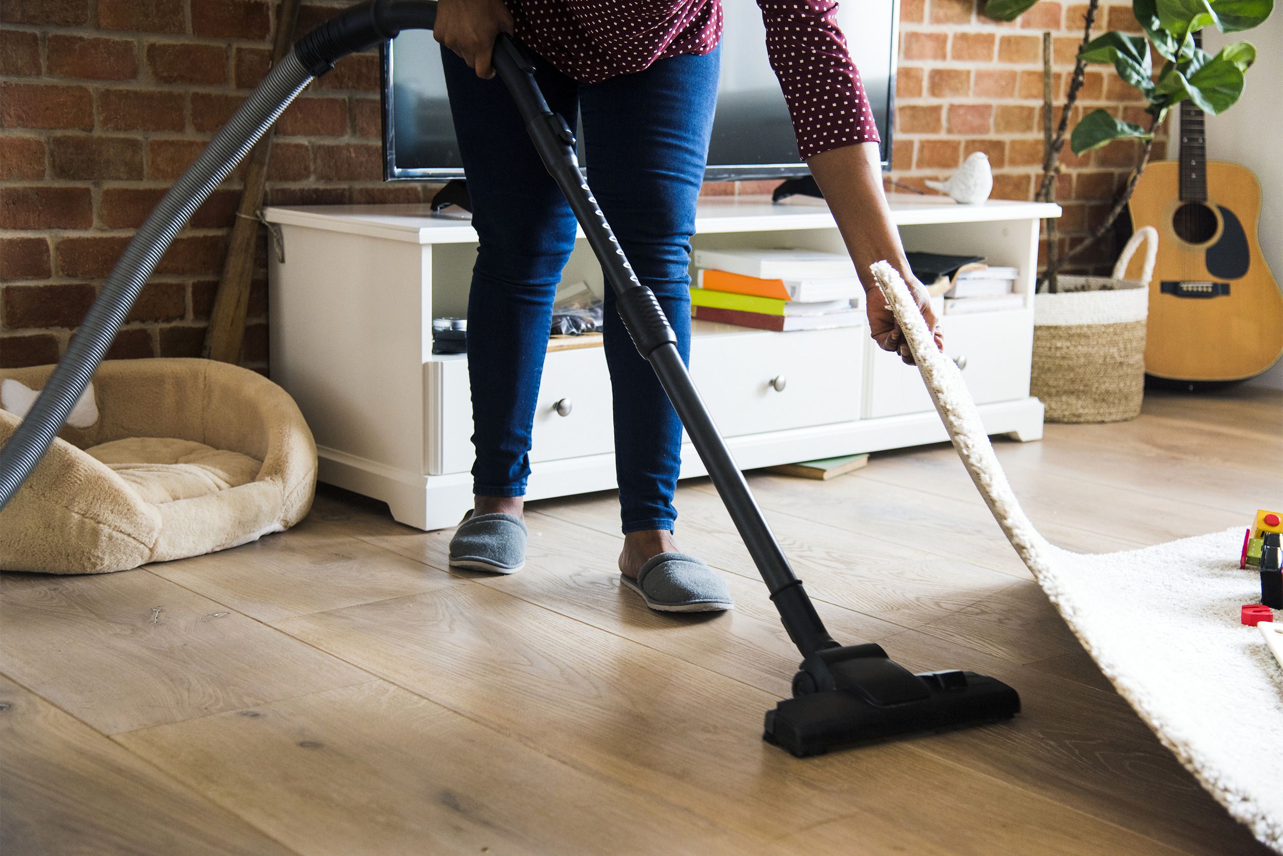 Mature woman lifting a rug while vacuuming floor in the living room with the TV and other items along a brick wall