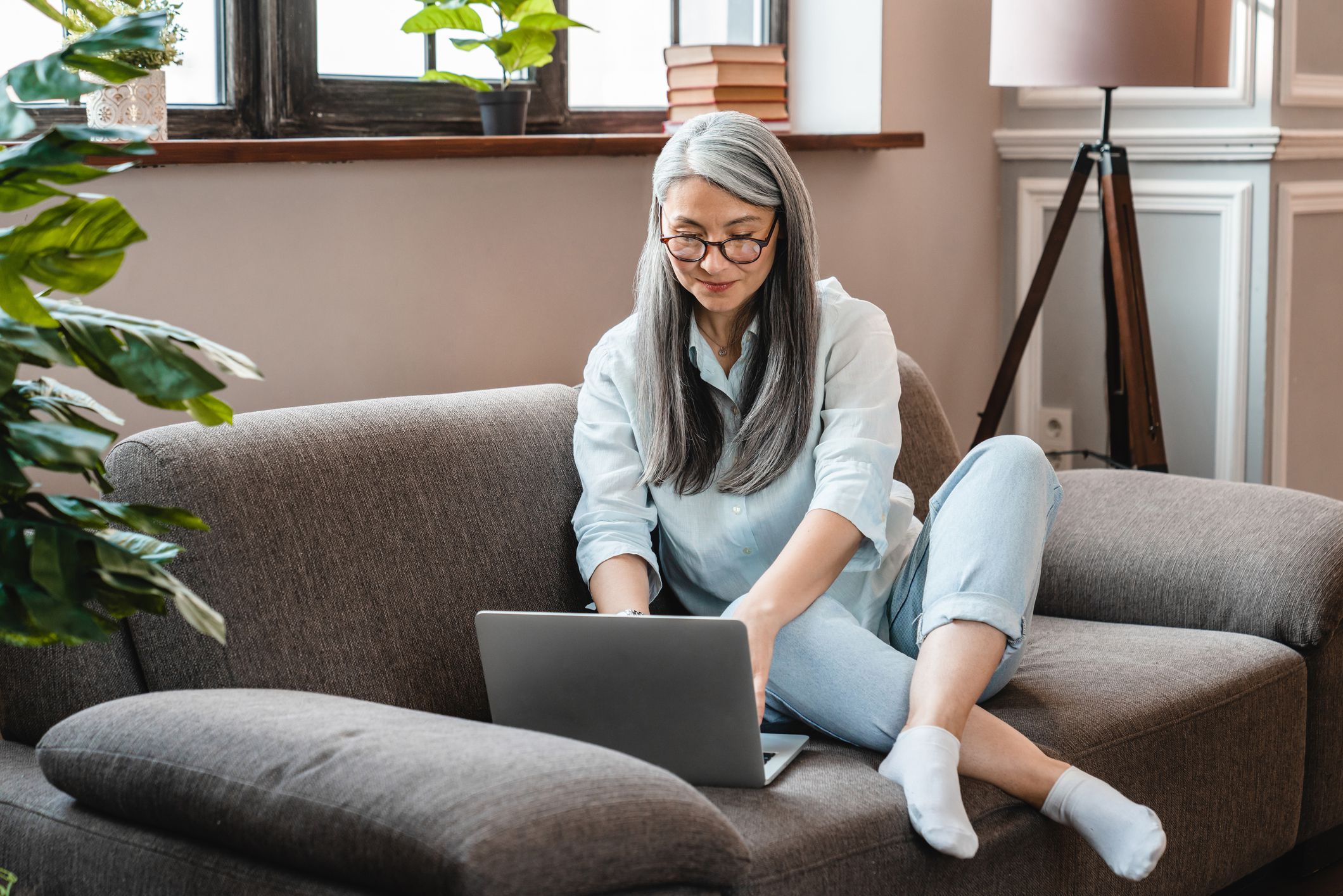 Mature woman using a laptop while sitting on a sofa in the living room of her home with plants and home furnishings in the background