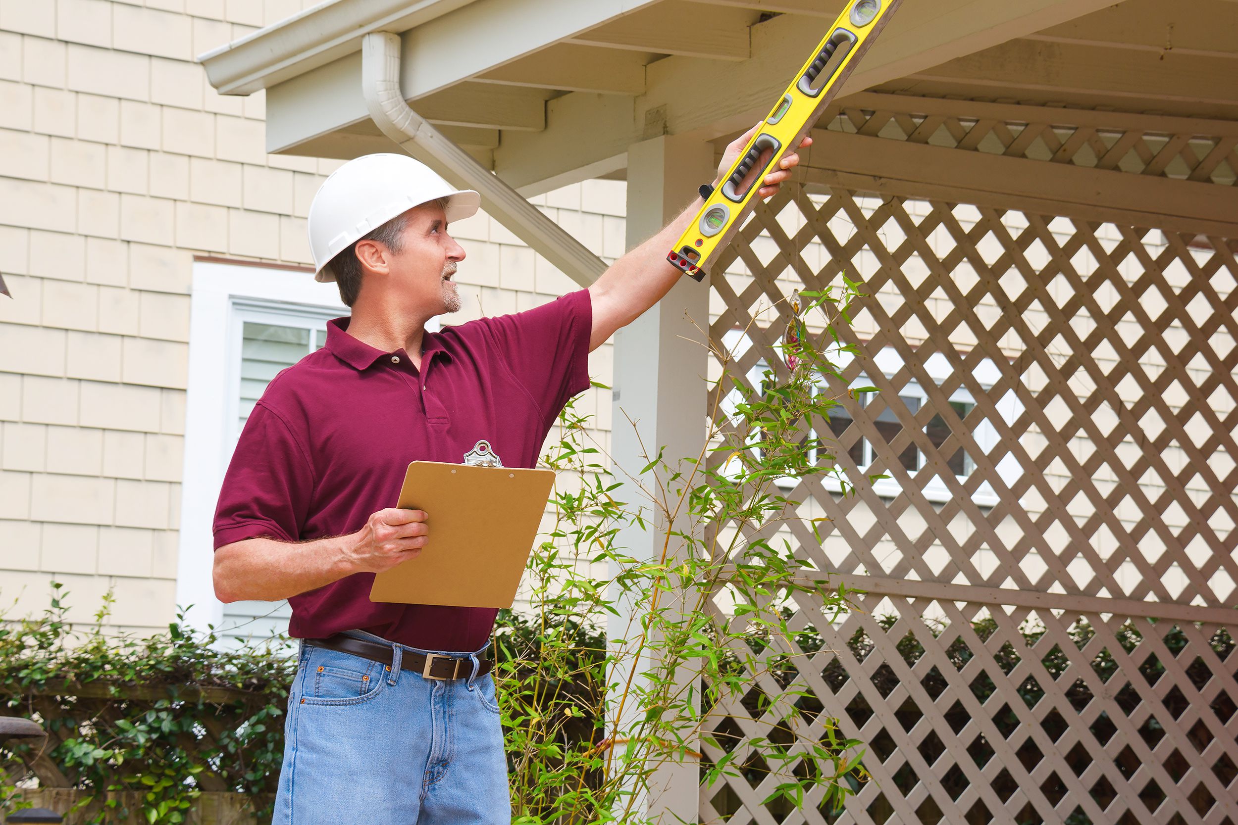 Senior home inspector in a hard hat holding a level and a clipboard outside a home doing an inspection
