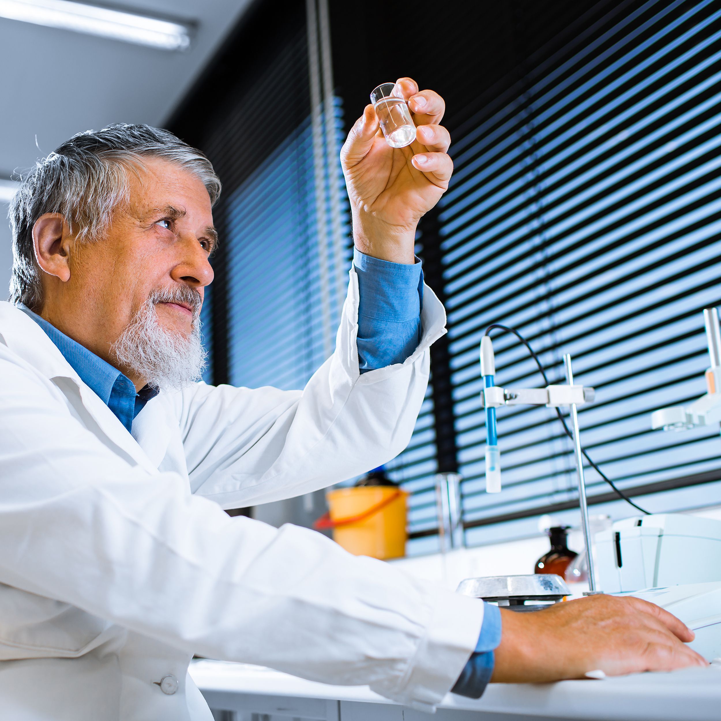 Senior male scientific researcher closely looking at a vial of liquid while holding it and sitting at a bench with items in the lab and windows