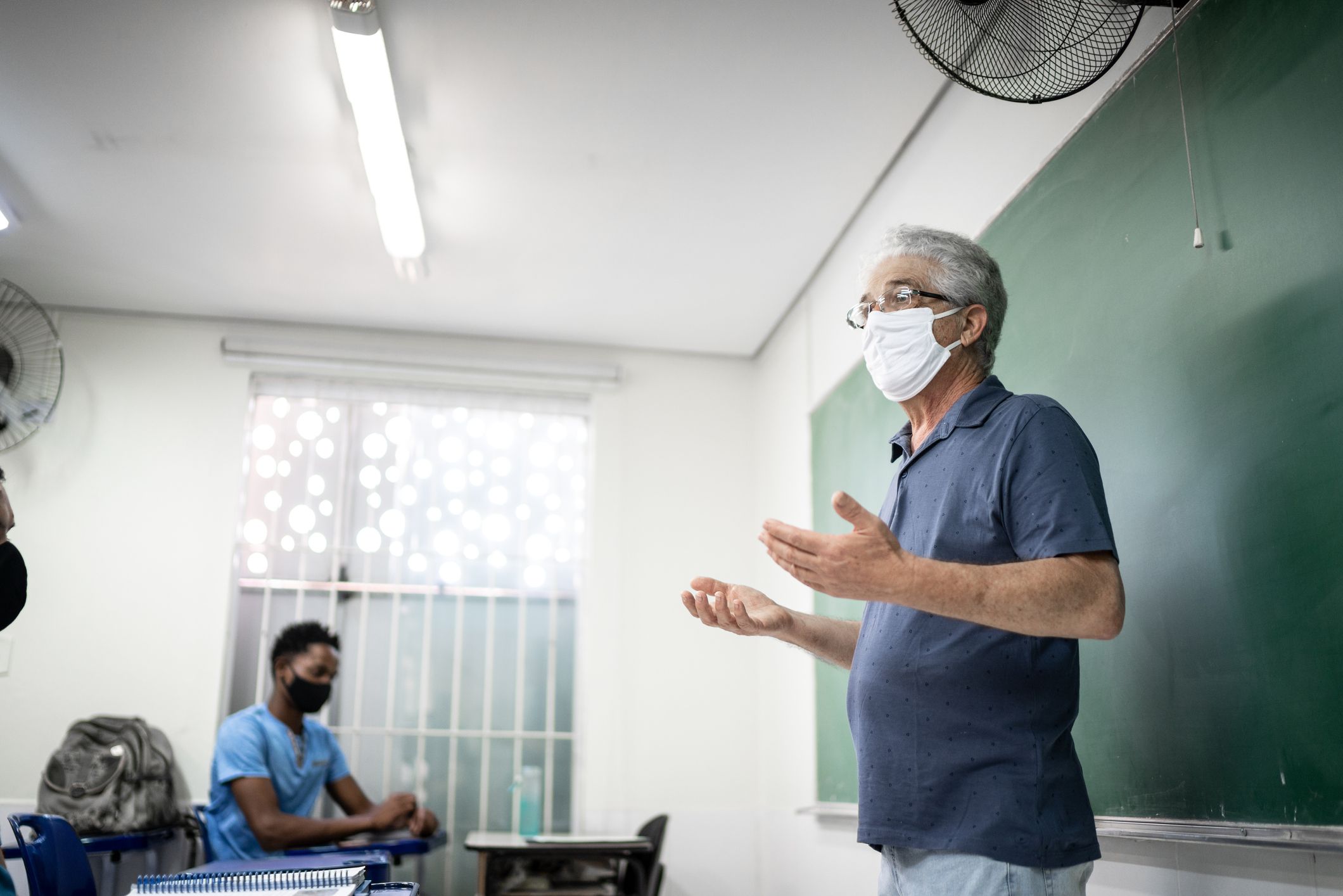 Senior male teacher with face mask teaching at an university, standing next to chalkboard with students and window in background
