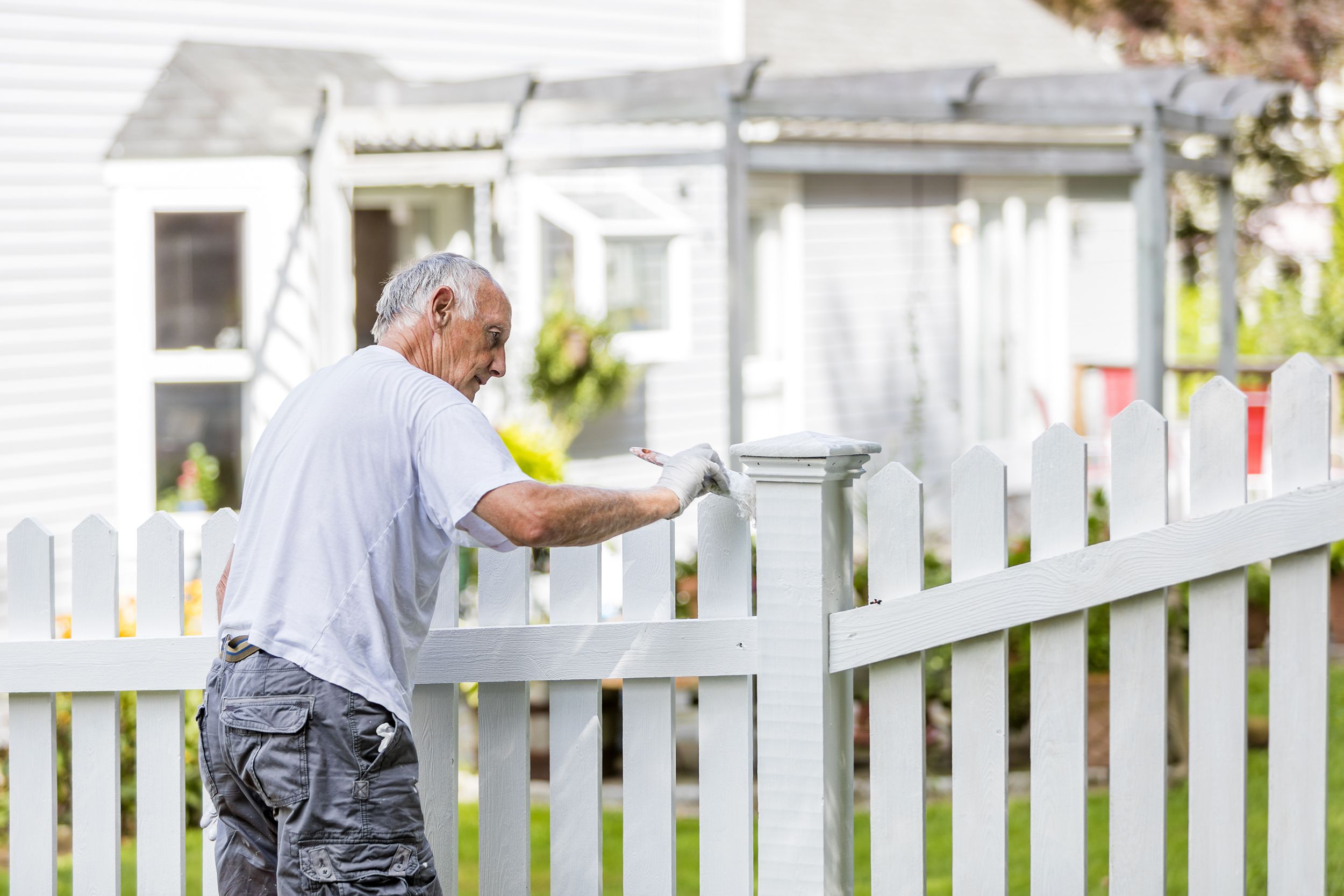 Senior man painting a white picket fence with a suburban home and backyard in the background