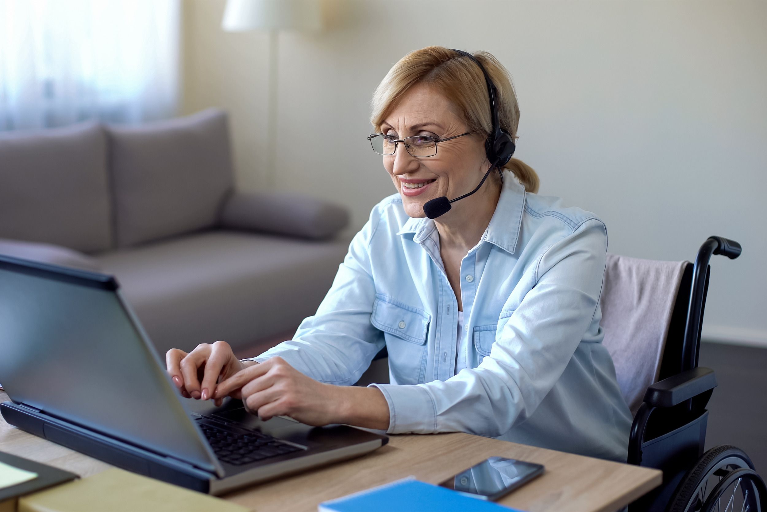 Senior woman sitting in a wheelchair, working on her laptop with a headset in her living room at home with a blurred background of living room