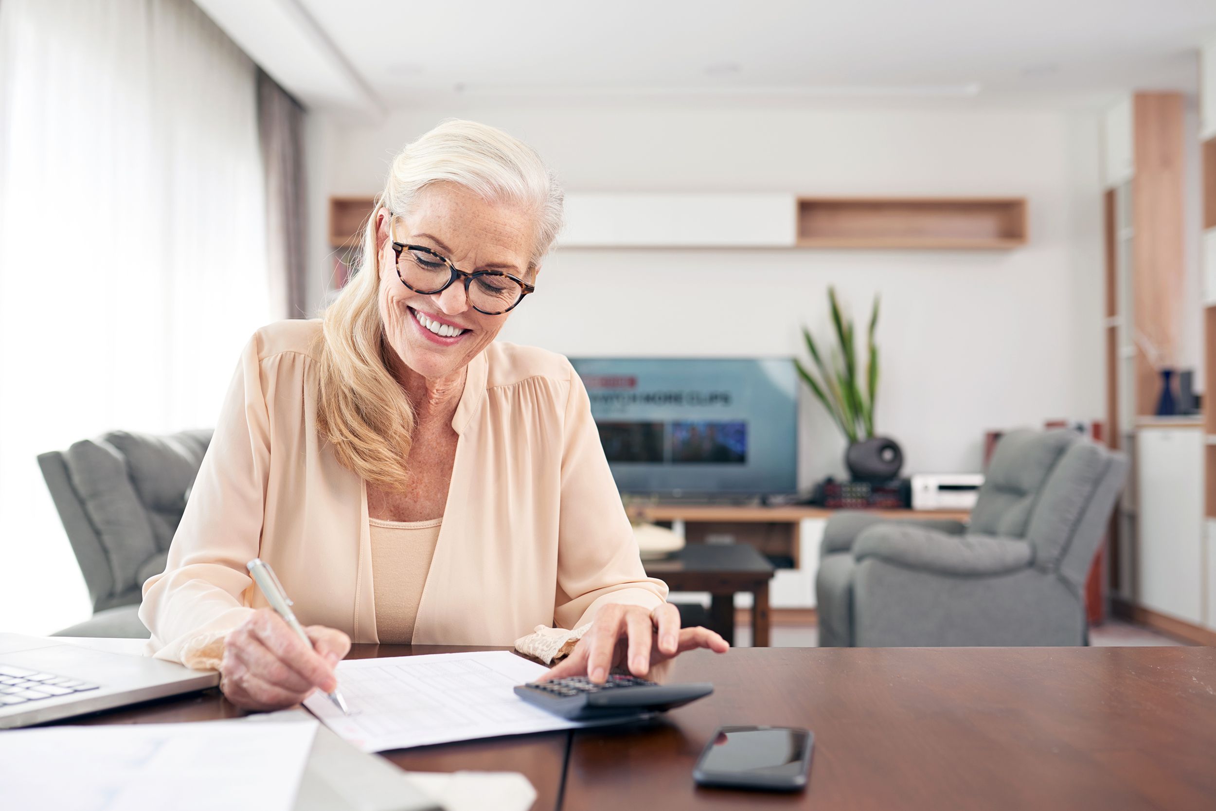 Smiling mature woman sitting at a desk in her living room, calculating expenses with living room in the background