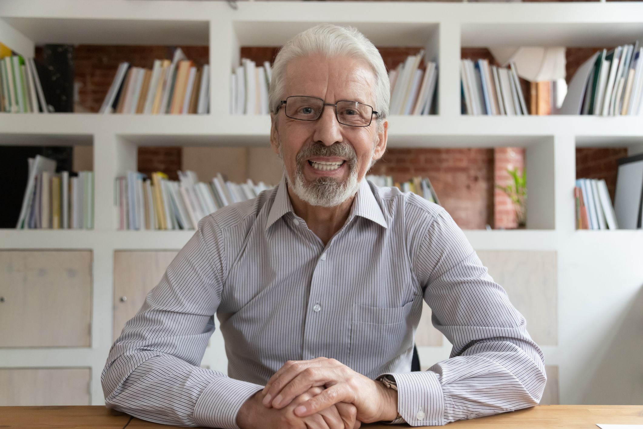 Smiling senior businessman with glasses sitting at desk, looking directly into webcam with bookcases in the background