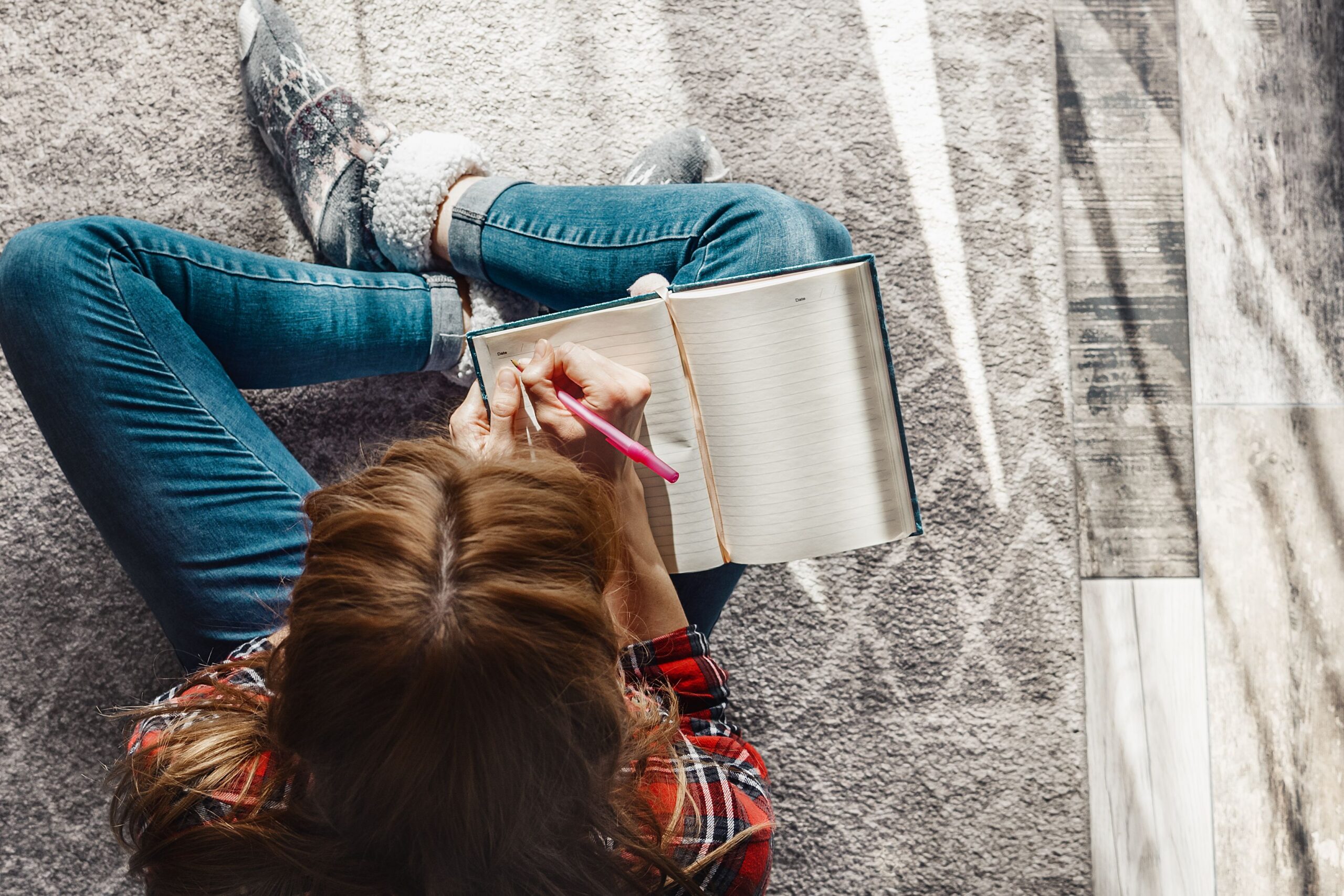 Top view of a mature woman sitting on a grey carpet on the floor, writing into a notebook with light coming through the window
